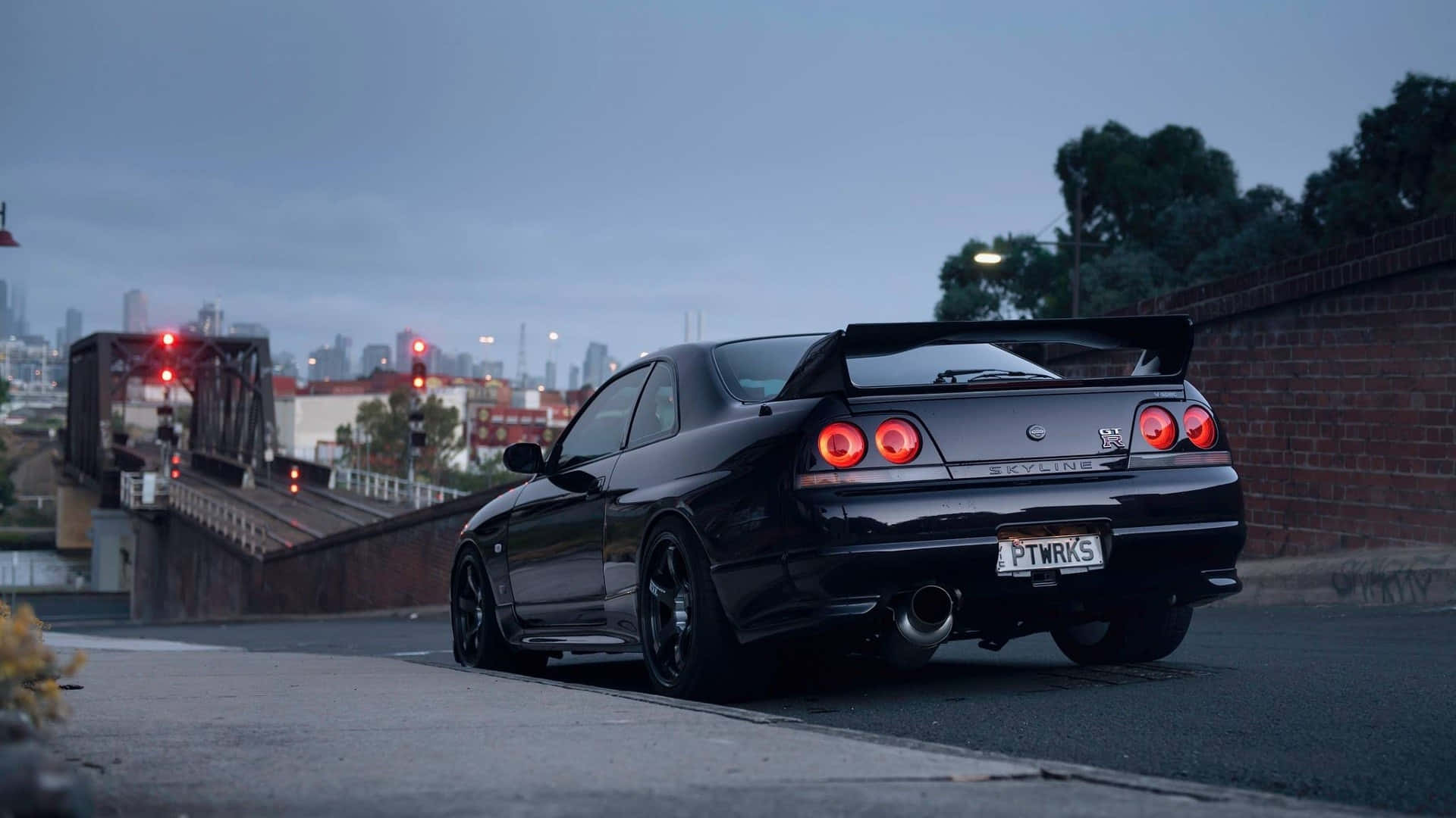 R33 GT-R 桌布 壁紙 — 一輛時尚的黑色日產Skyline GT-R在黃昏時停泊在橋上，帶有發光的紅色尾燈和城市天際線背景。