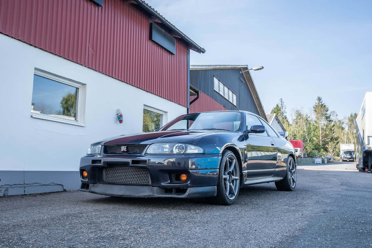 R33 GT-R 桌布 壁紙 — 一輛時尚的黑色日產Skyline GT-R停在紅白相間的工業建築前，藍天白雲。