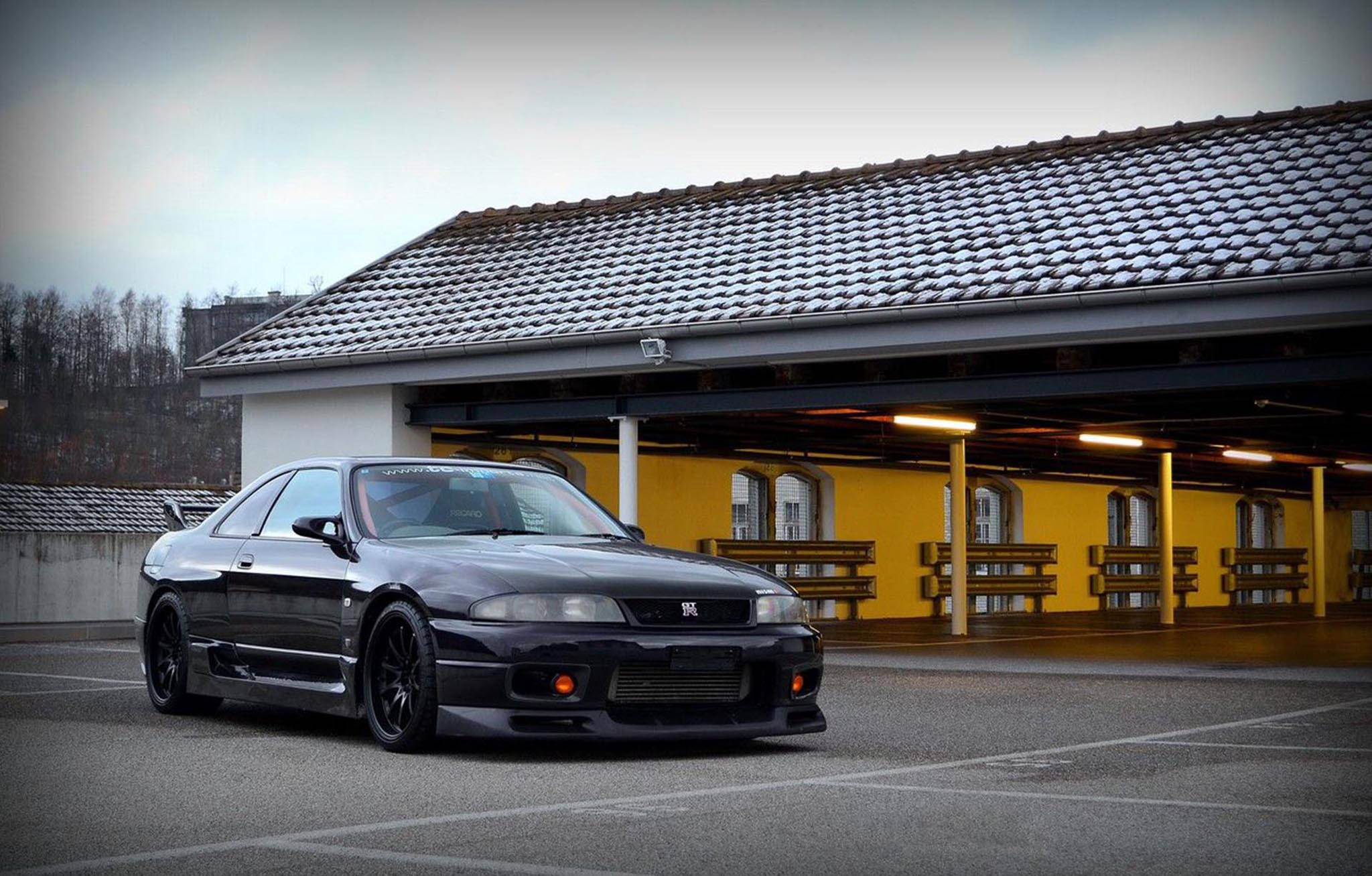 Gtr 桌布 壁紙 — 一輛時尚的黑色日產Skyline GT-R停放在黃色瓦頂建築物前，天空多雲。