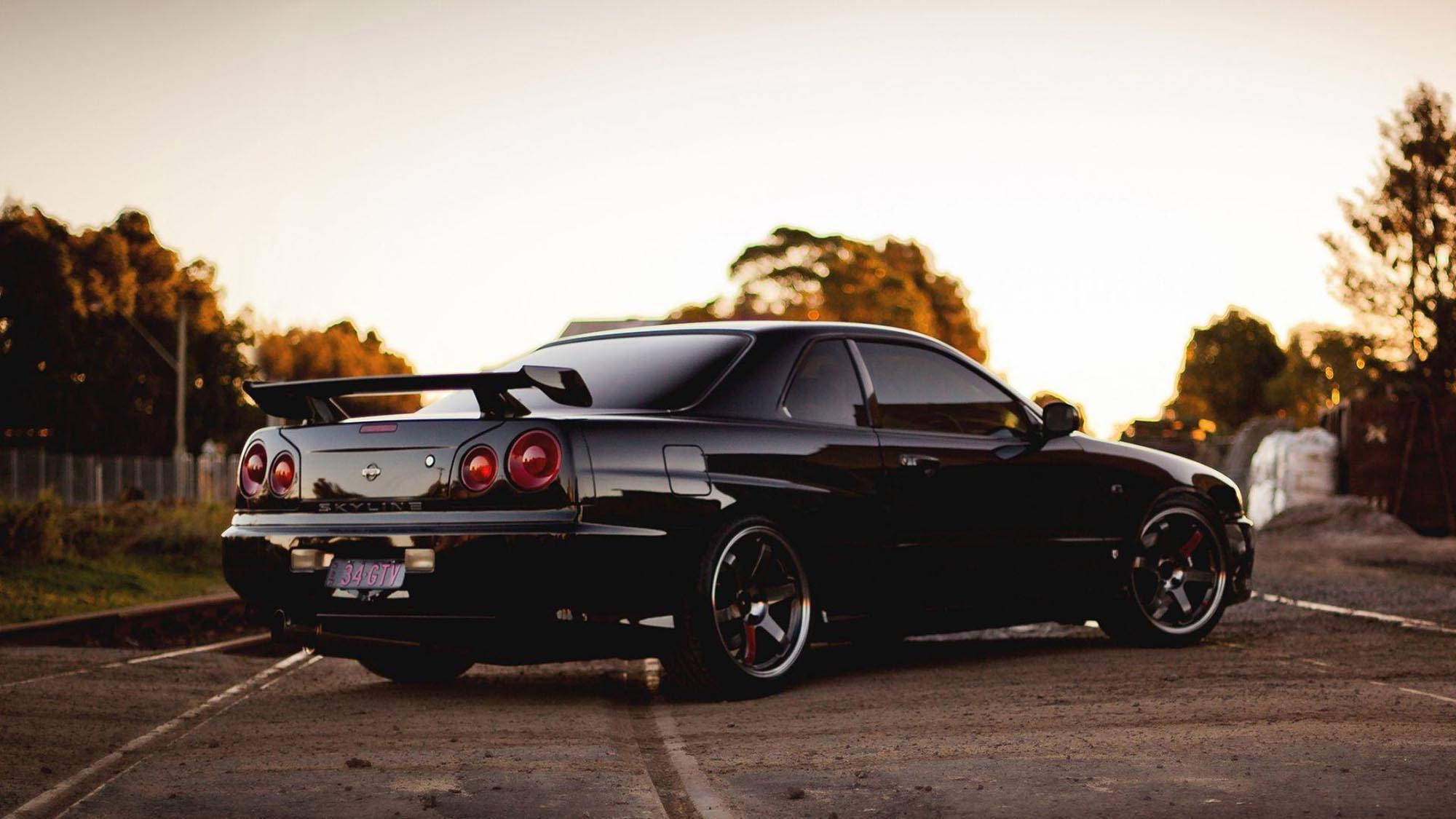 Gtr 桌布 壁紙 — 一輛時尚的黑色日產Skyline R34跑車停放在鋪砌的停車場，背景有樹木，正值黃金時段。