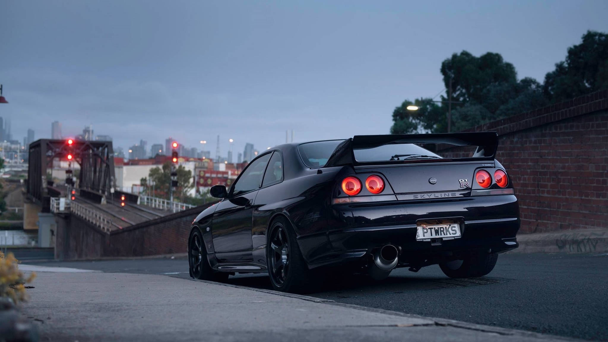 Gtr 桌布 壁紙 — 一輛時尚的黑色日產Skyline GT-R在黃昏時停泊在橋上，帶有發光的紅色尾燈和城市天際線背景。