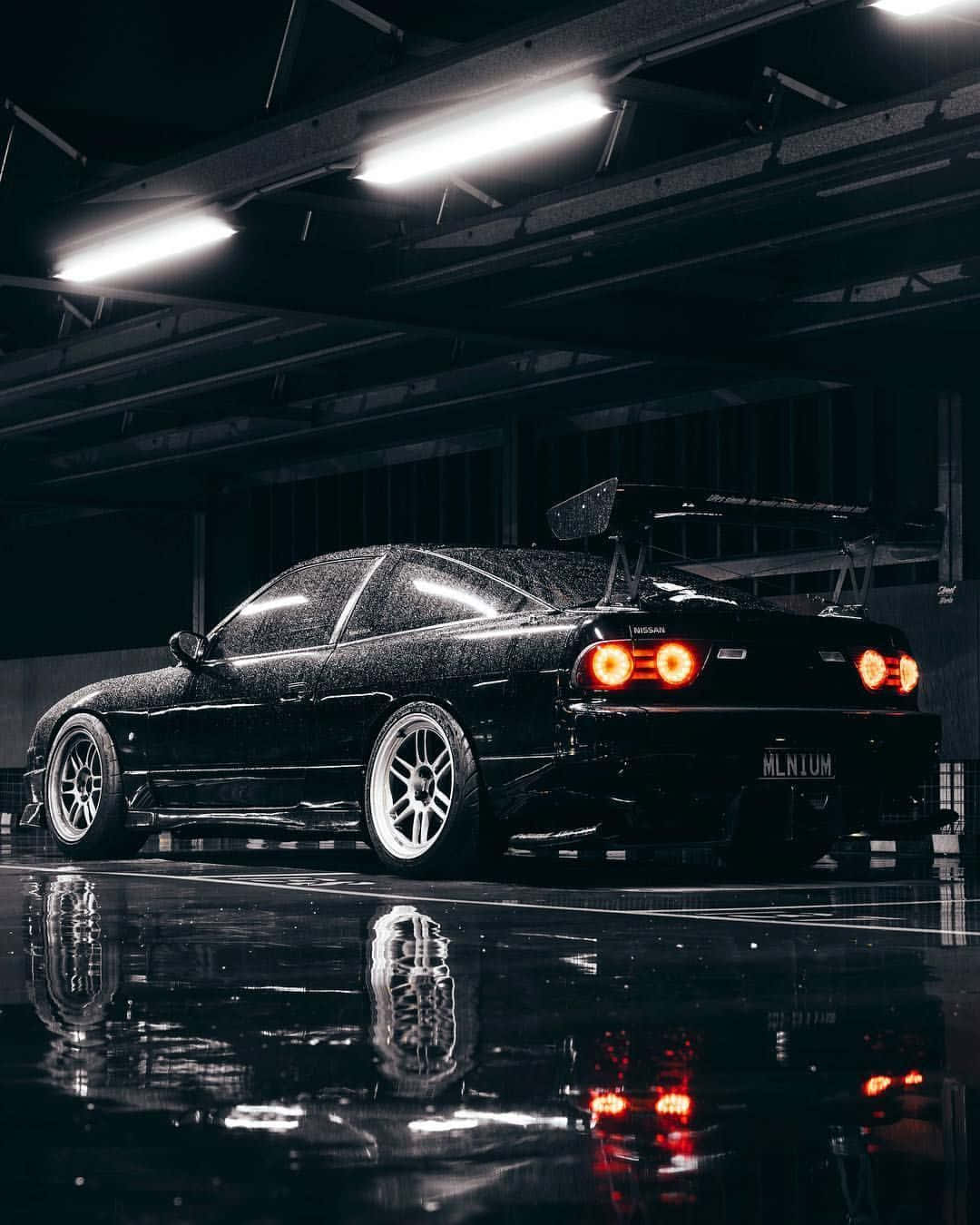 JDM iPhone 桌布 壁紙 — 一輛時尚的黑色日產跑車，帶有發光的紅色尾燈，停在潮濕的地下車庫中，在螢光燈下反射出其銳利的線條。