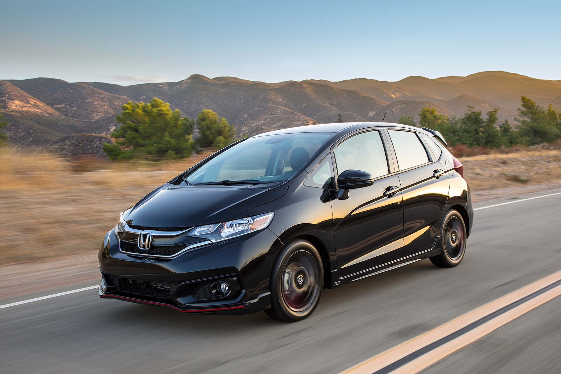 Honda Fit 桌布 壁紙 — 一輛時尚的黑色本田Fit在沙漠公路上高速行駛，背景是陽光普照的山脈和稀疏的綠植，呈現動態模糊效果。