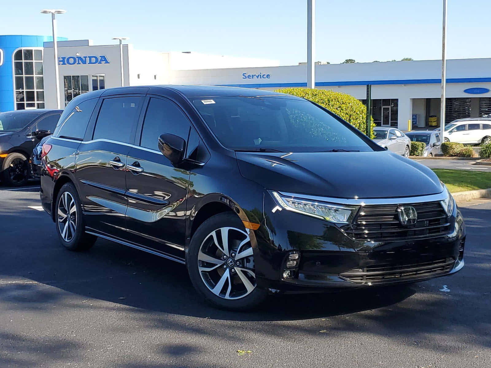 Honda Odyssey 桌布 壁紙 — 一輛時尚的黑色本田小型客車在明亮的陽光下，於經銷商場地中閃閃發光，背景有其他汽車和服務大樓。