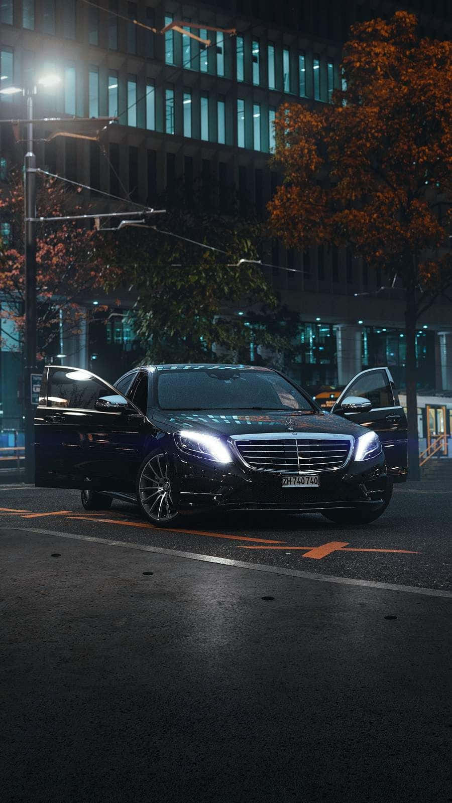Mercedes Benz Clase E 桌布 壁紙 — 一輛時尚的黑色梅賽德斯轎車，車頭燈閃爍，停在潮濕的城市街道上，夜晚路燈下，背景有秋樹和亮燈的建築物。