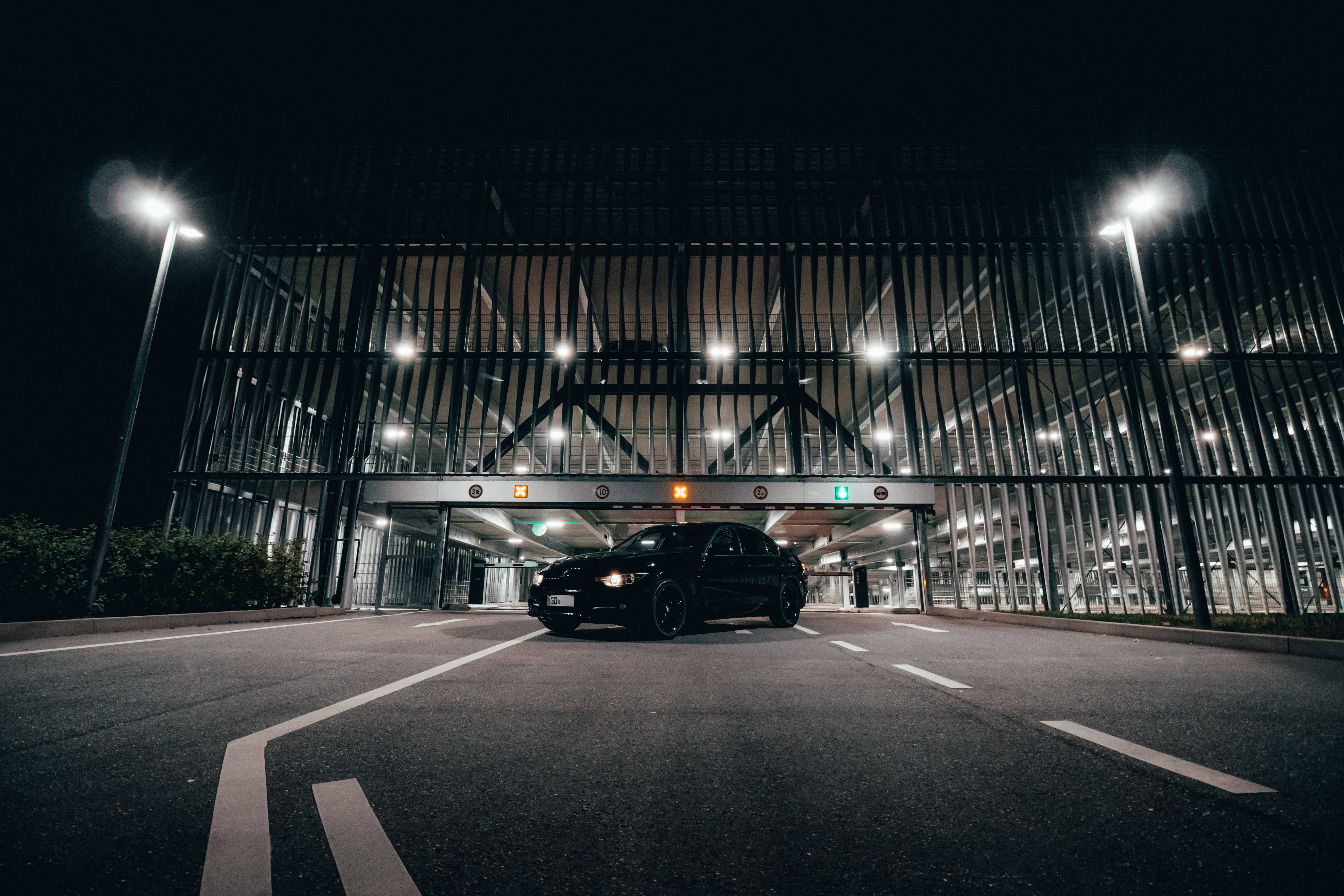 m 桌布 壁紙 — 一輛時尚的黑色汽車在夜間停放在現代金屬結構下，明亮的街燈投下戲劇性的陰影。