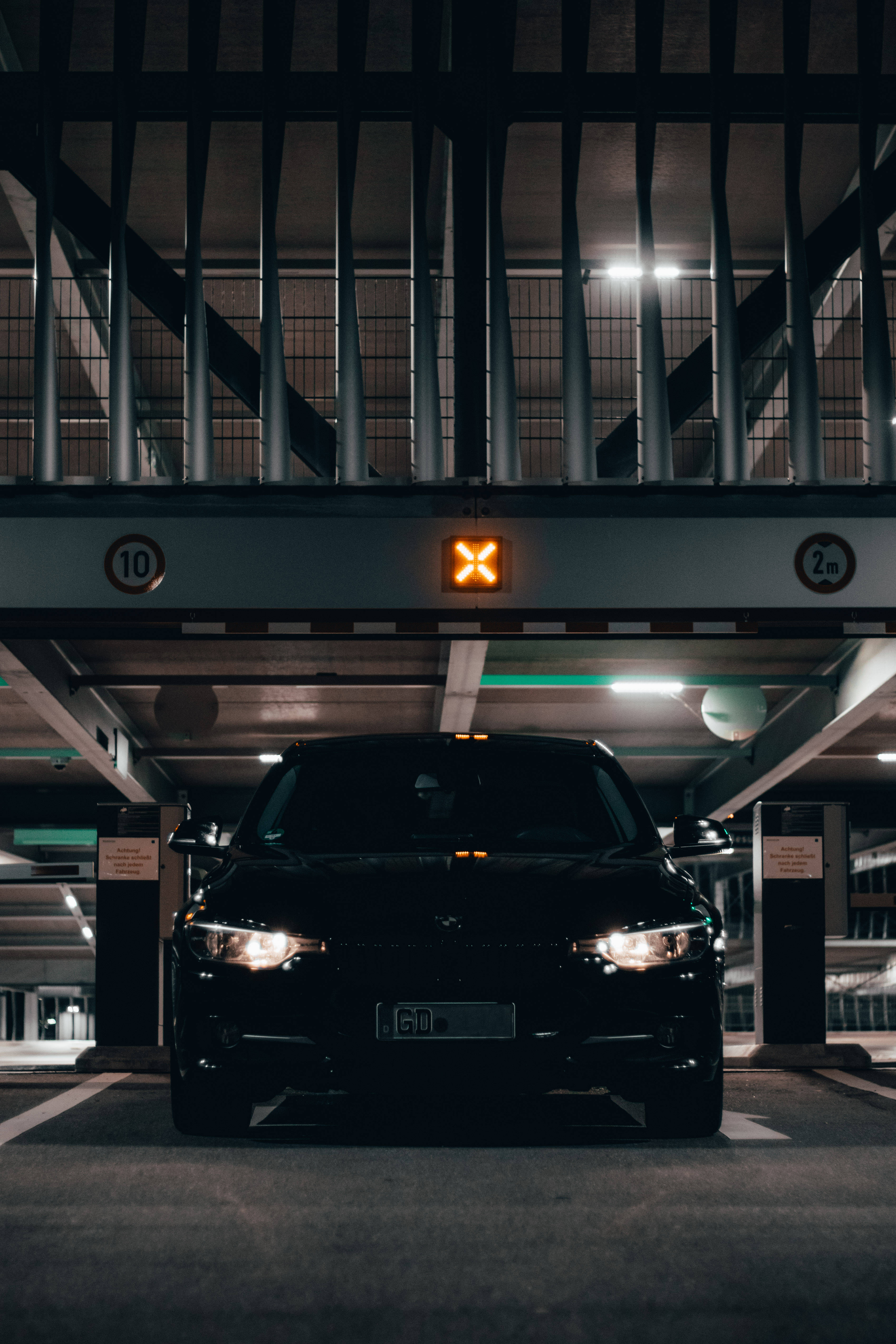 m 桌布 壁紙 — 一輛時尚的黑色汽車在燈光昏暗的停車場內向前，上方有一個發光的橙色「X」標誌。
