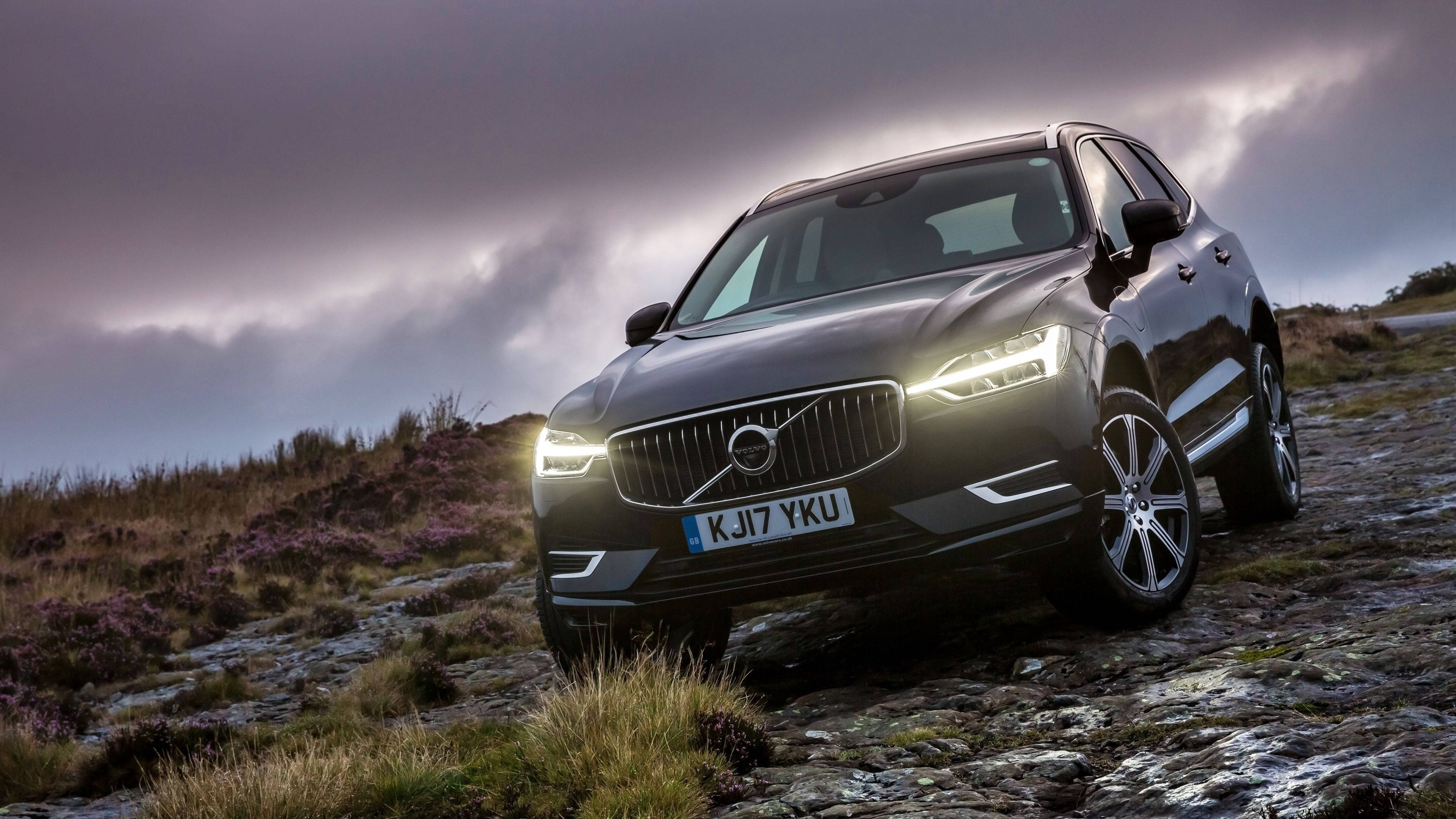 Volvo 桌布 壁紙 — 一輛時尚的黑色沃爾沃SUV，車頭燈閃閃發光，停放在崎嶇的、佈滿紫色石楠花的山坡上，背景是陰沉的暮色天空。