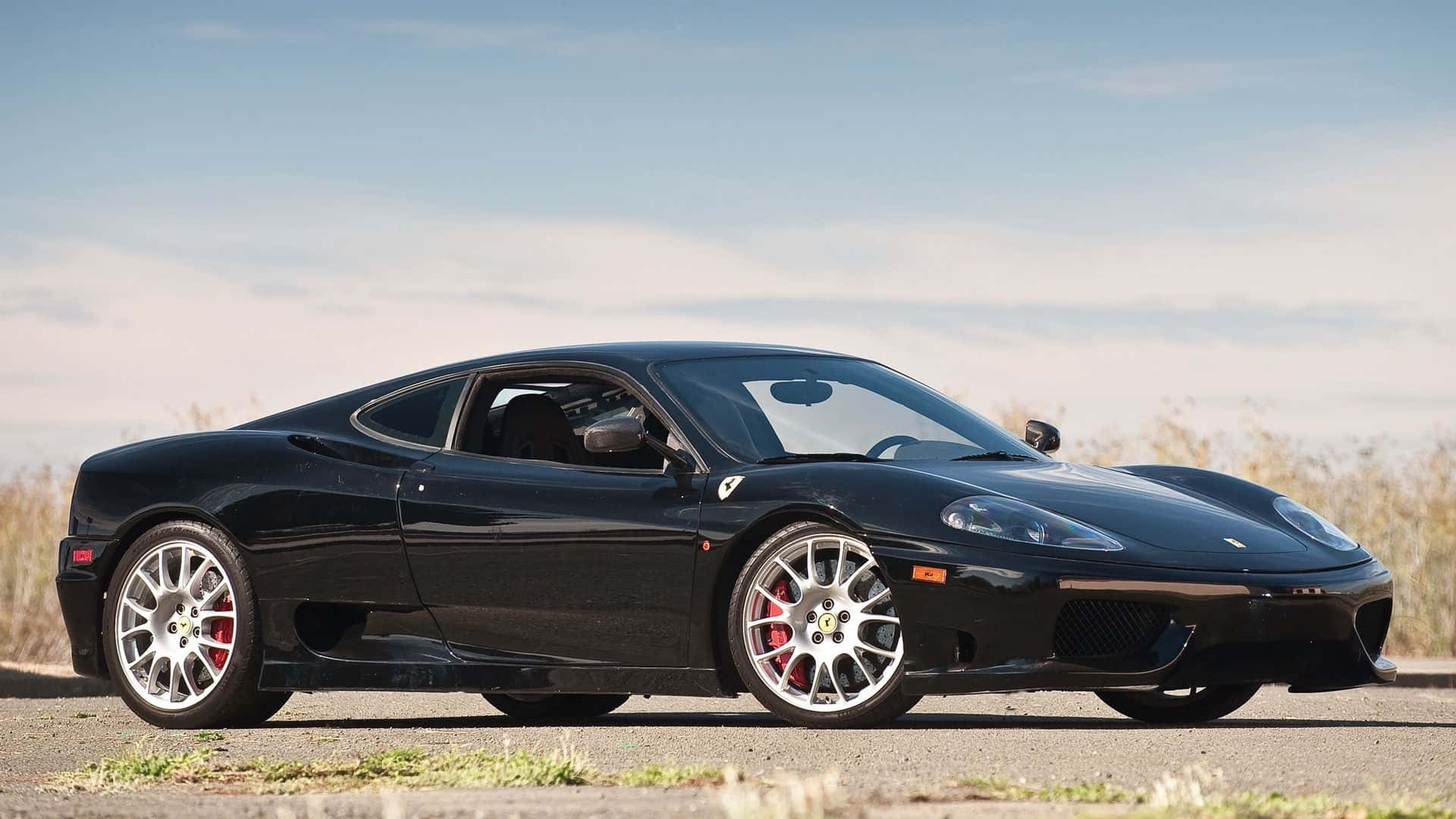 Ferrari 360 Modena 桌布 壁紙 — 一輛時尚的黑色法拉利 360 跑車停放在鋪砌的道路上，背景有乾草和晴朗的藍天。