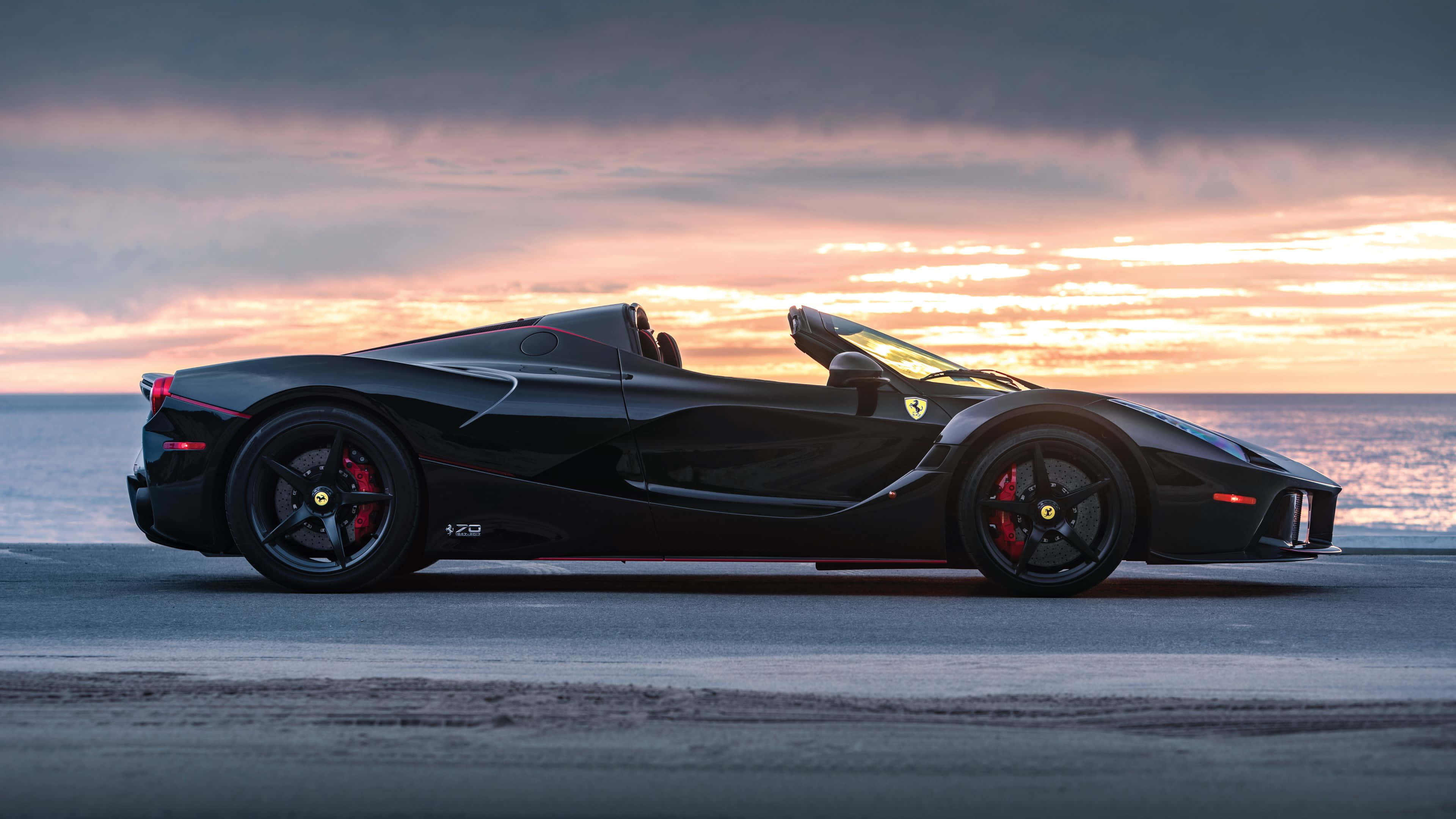 Vintage Ferrari 桌布 壁紙 — 一輛時尚的黑色法拉利 LaFerrari Aperta 停泊在日落時分的海岸公路上，海洋上空有鮮豔的橙色和粉紅色雲彩。