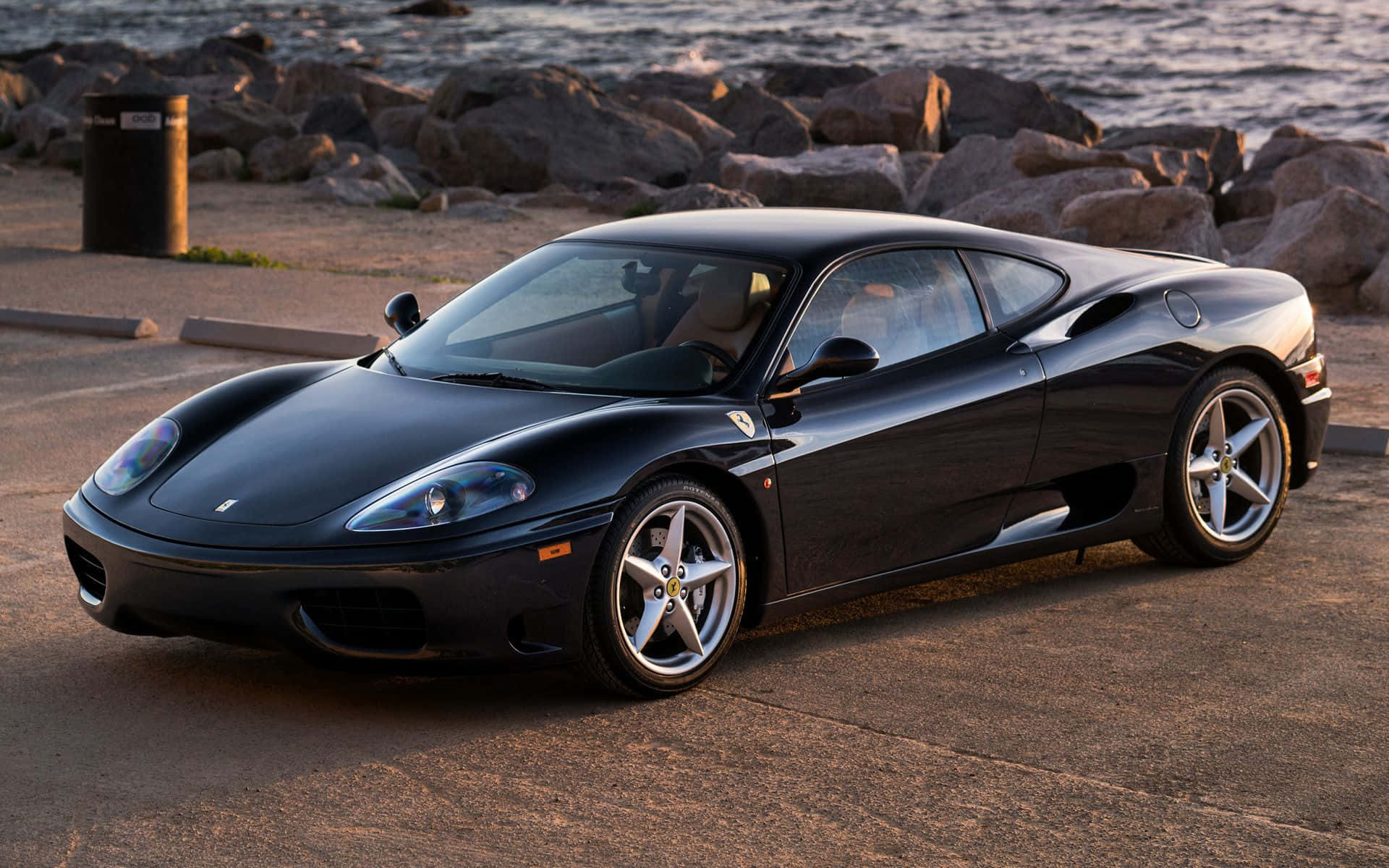Ferrari 360 Modena 桌布 壁紙 — 一輛時尚的黑色法拉利360跑車停泊在黃金時段岩石海岸線旁的鋪砌停車場上。