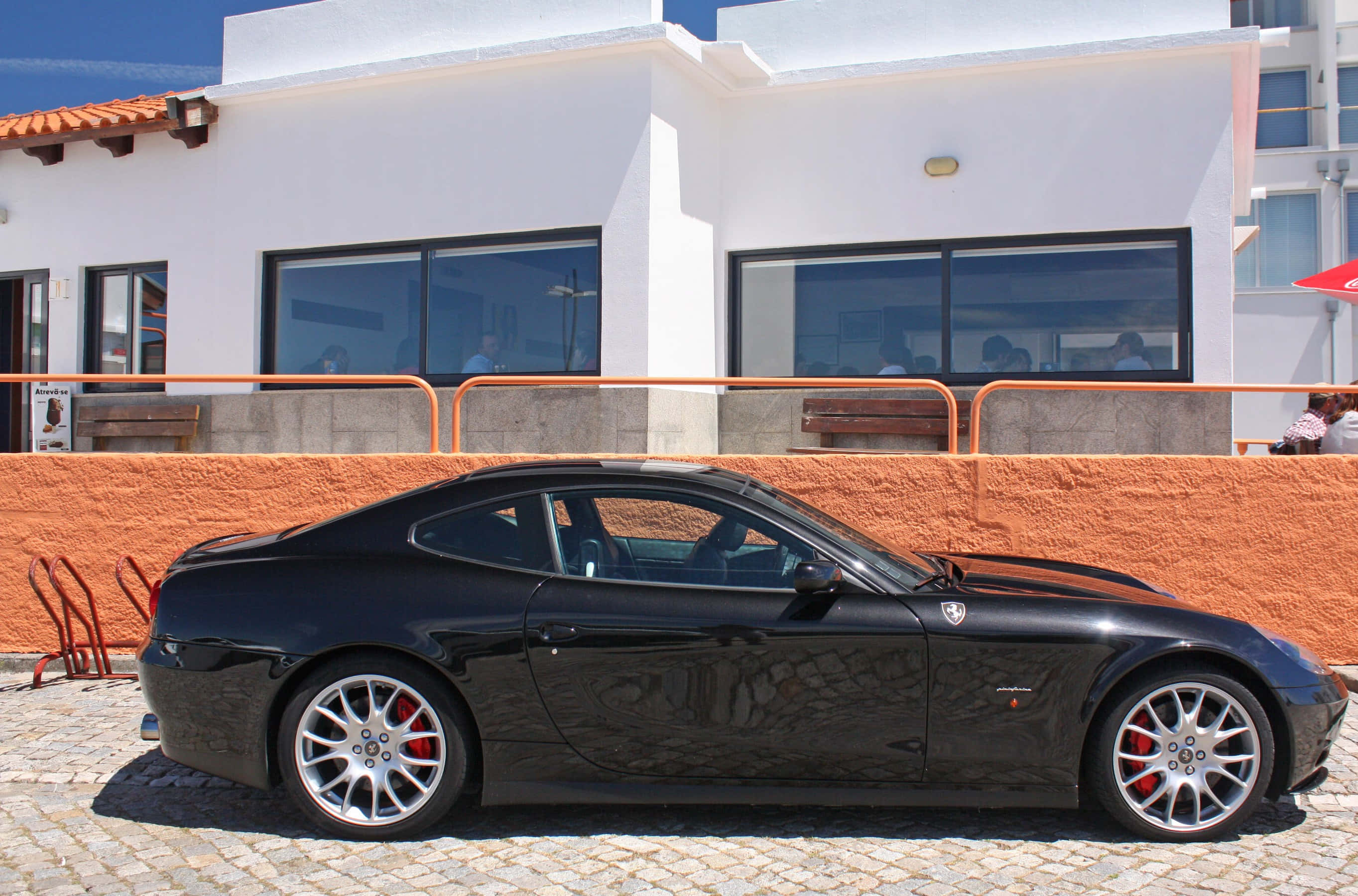 Ferrari 612 Scaglietti 桌布 壁紙 — 一輛時尚的黑色法拉利612 Superamerica停泊在鵝卵石路上，前方是白色建築物，牆壁為橙色，陽光普照。