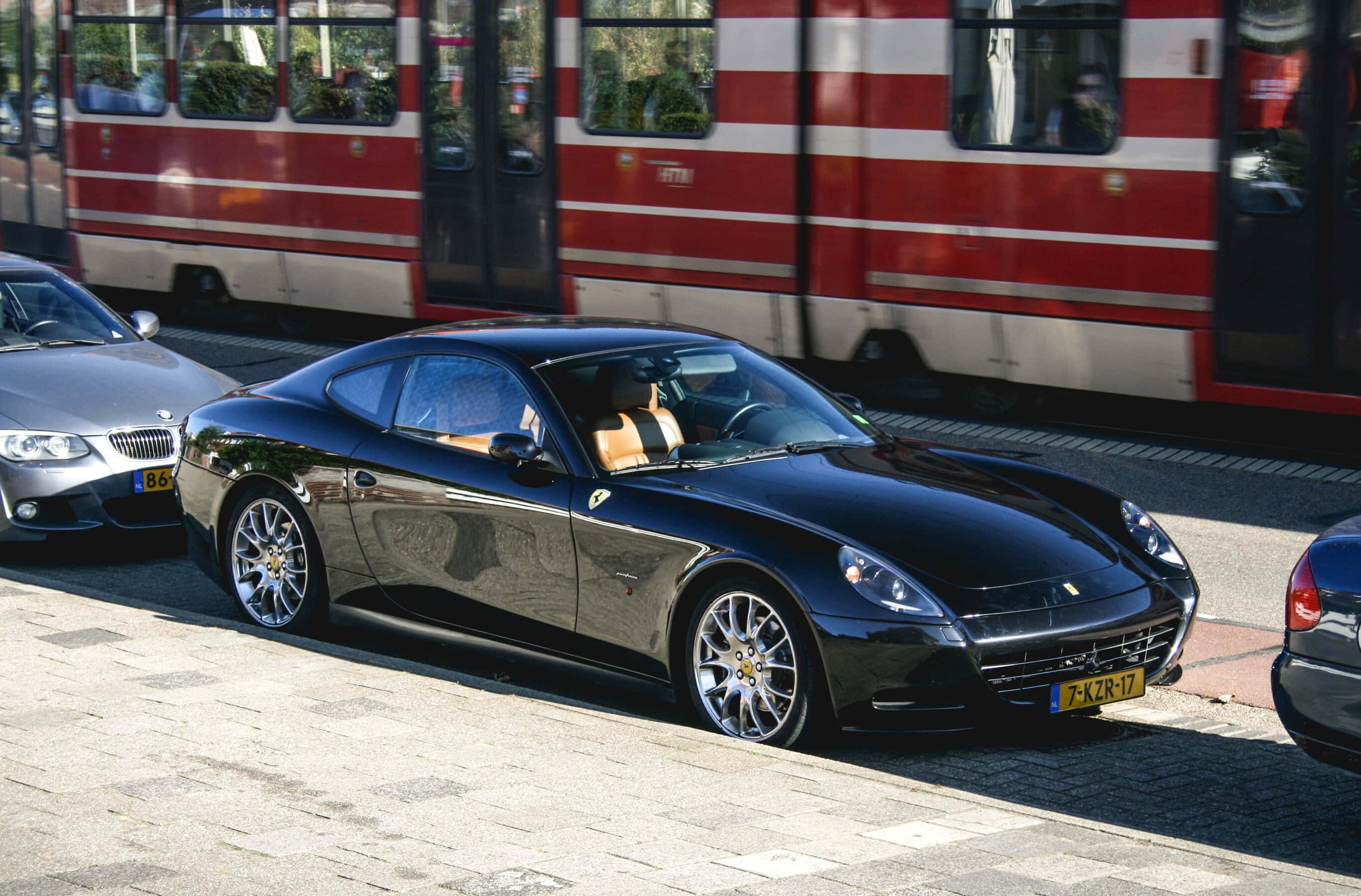 Ferrari 612 Scaglietti 桌布 壁紙 — 一輛時尚的黑色法拉利California停泊在陽光普照的鵝卵石街道上，旁邊是一列紅白相間的火車。