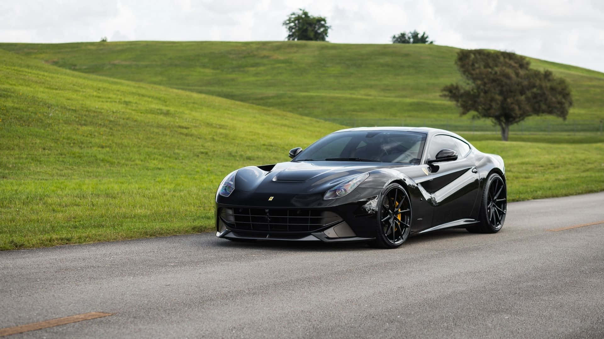 Ferrari F12 Berlinetta 桌布 壁紙 — 一輛時尚的黑色法拉利F12停泊在蜿蜒的道路旁，旁邊是起伏的綠色山丘，天空多雲。