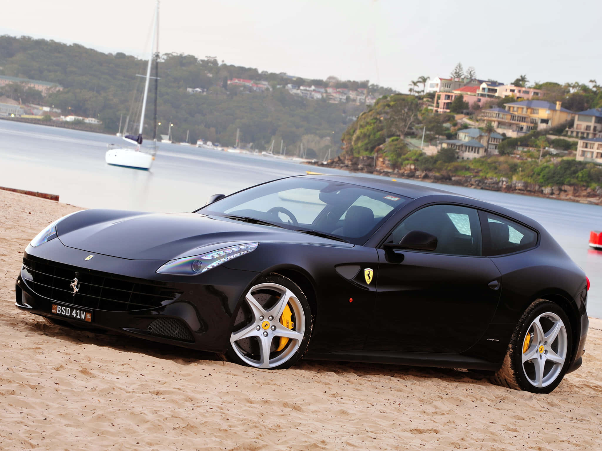 Ferrari Ff 桌布 壁紙 — 一輛時尚的黑色法拉利FF停在沙灘上，背景有帆船和沿海房屋。