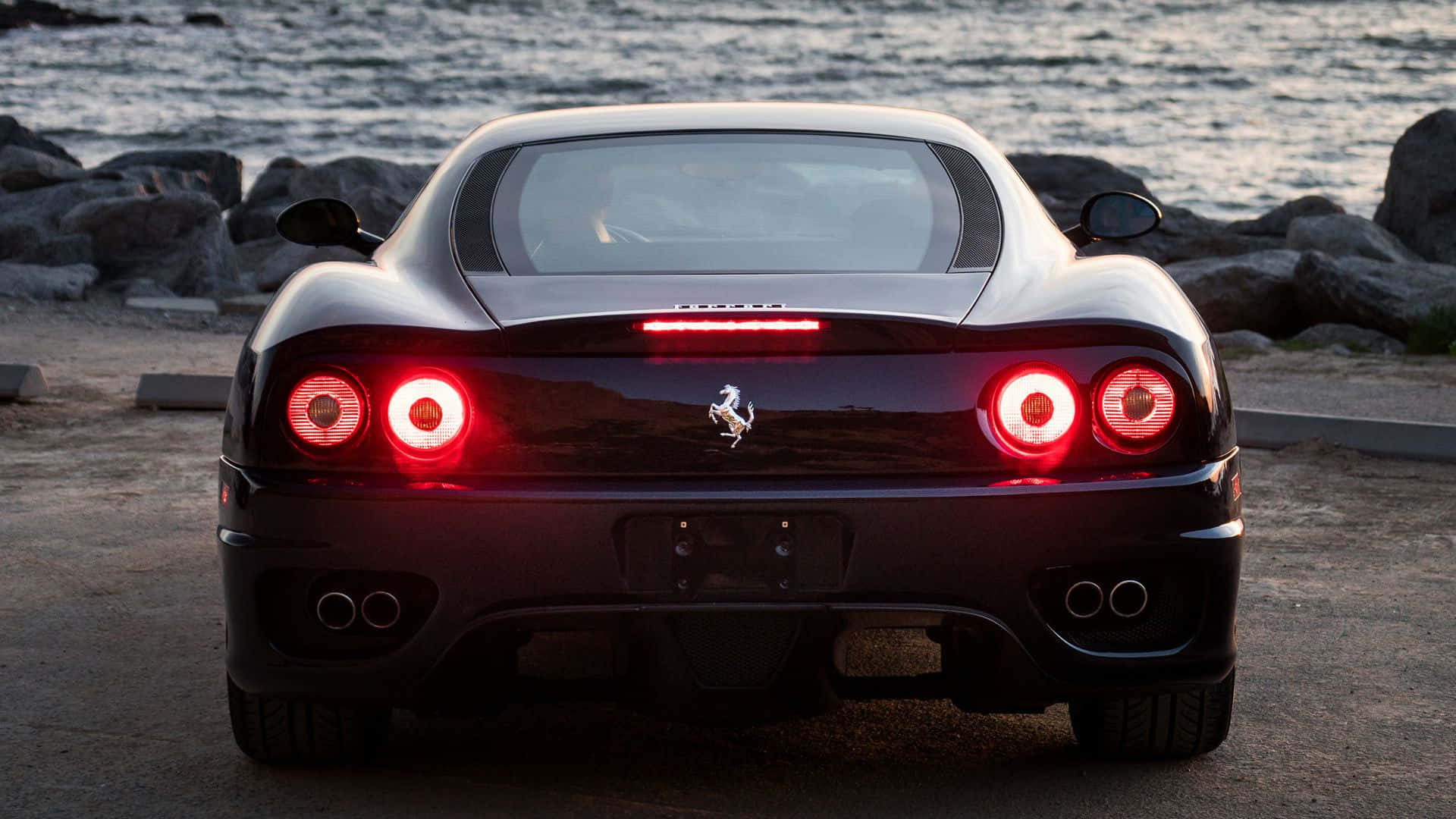 Ferrari 360 Modena 桌布 壁紙 — 一輛時尚的黑色法拉利跑車，帶有發光的紅色尾燈，在黃昏時停泊在岩石海濱旁。