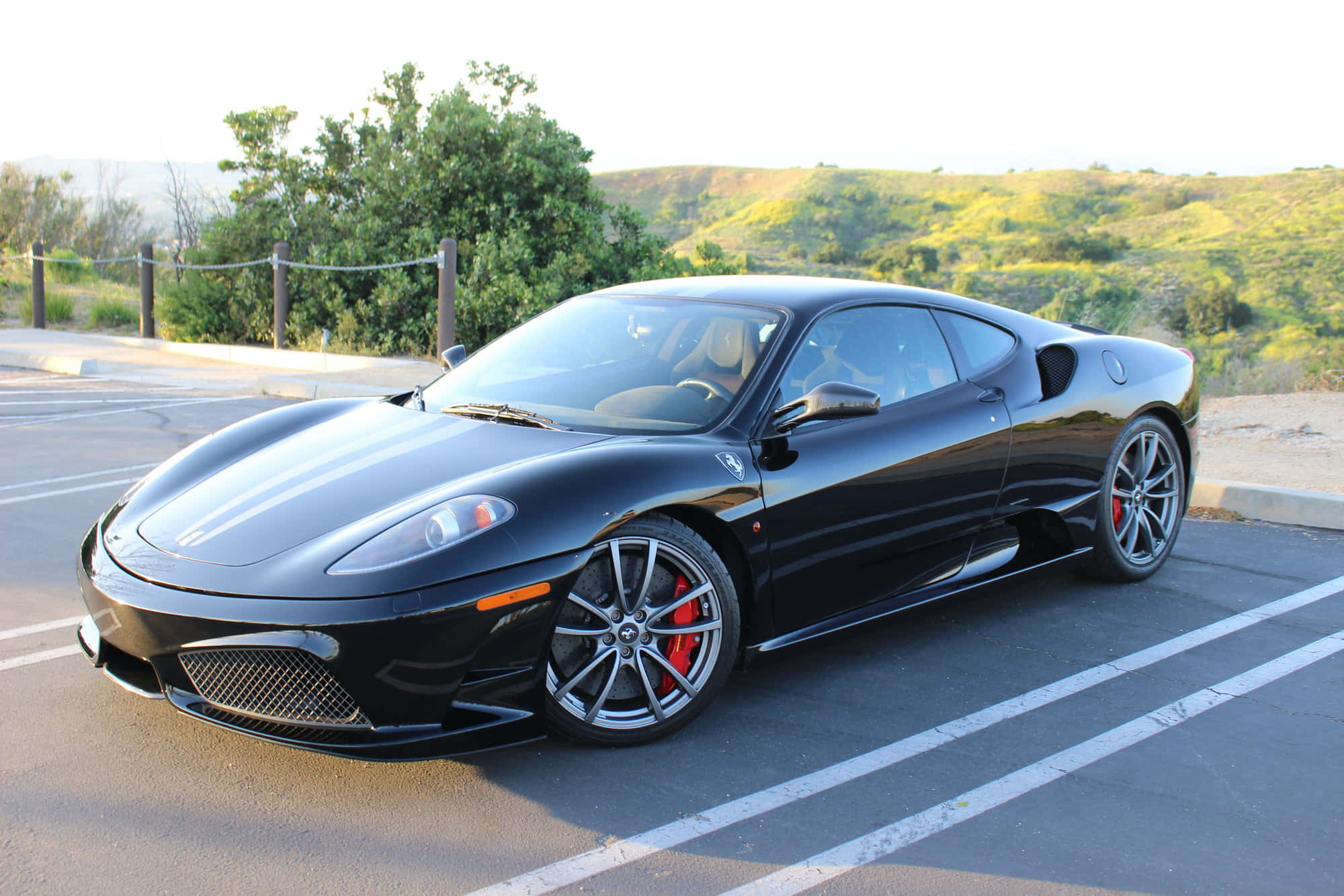 Ferrari F430 桌布 壁紙 — 一輛時尚的黑色法拉利跑車，配備紅色制動卡鉗，停放在陽光普照的停車場，背景是起伏的綠色山丘。