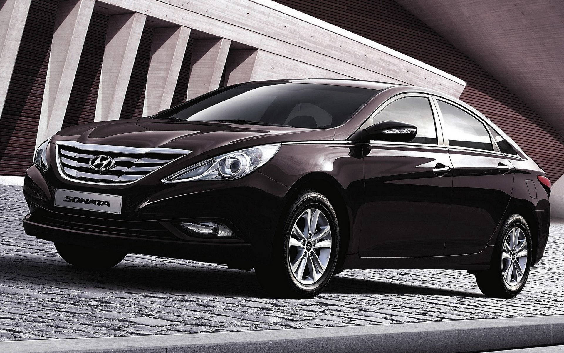 Hyundai 桌布 壁紙 — 一輛時尚的黑色現代索納塔轎車在明亮的燈光下，在現代混凝土建築旁邊的鵝卵石街道上閃閃發光。