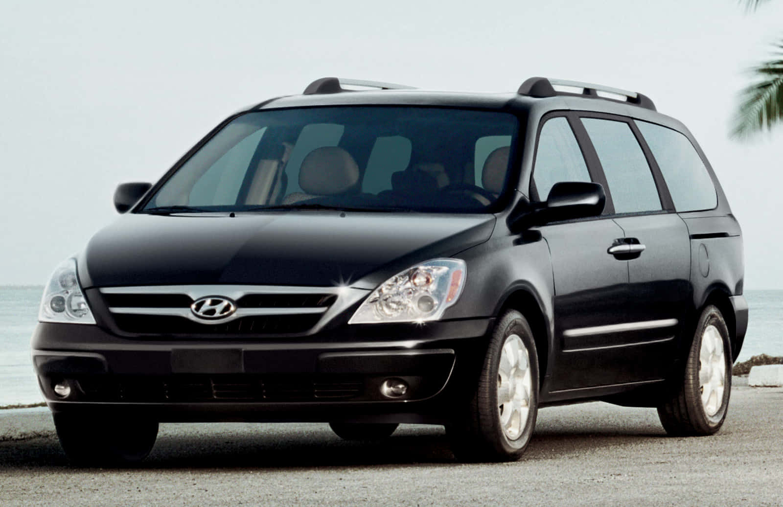 Hyundai Entourage 桌布 壁紙 — 一輛時尚的黑色現代迷你廂型車停放在沿海的碎石路上，背景是海洋和棕櫚葉。