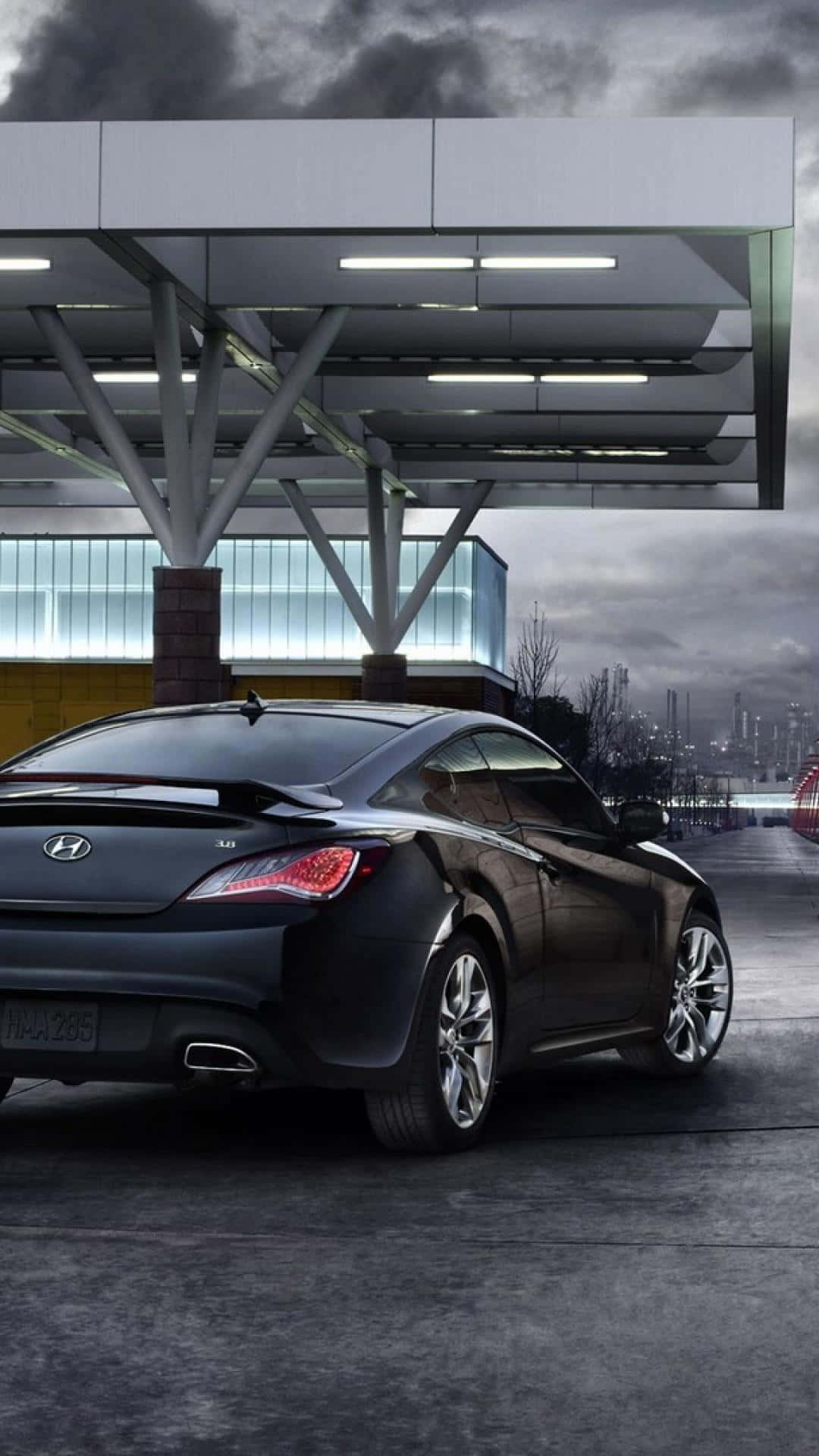 Hyundai Genesis 桌布 壁紙 — 一輛時尚的黑色現代運動轎跑車停放在現代加油站頂棚下，背景是陰沉的工業天際線。