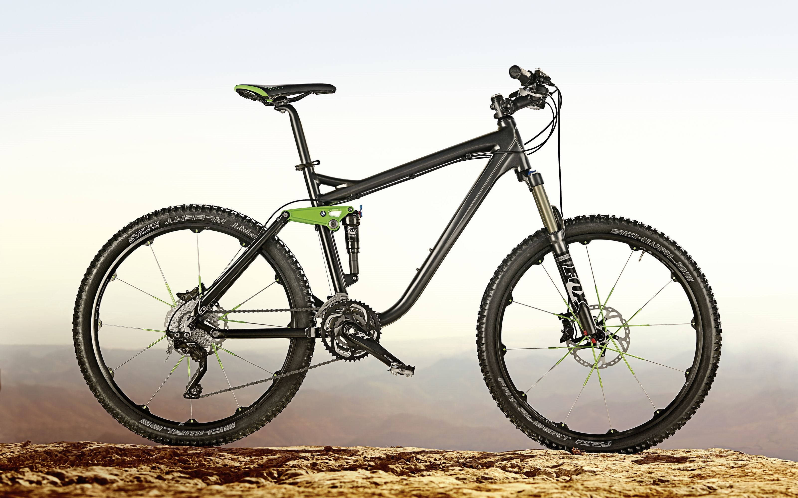 Mountain Bike 桌布 壁紙 — 一輛時尚的黑色登山自行車，帶有萊姆綠色點綴，矗立在岩石懸崖上，俯瞰著朦朧的沙漠山谷。