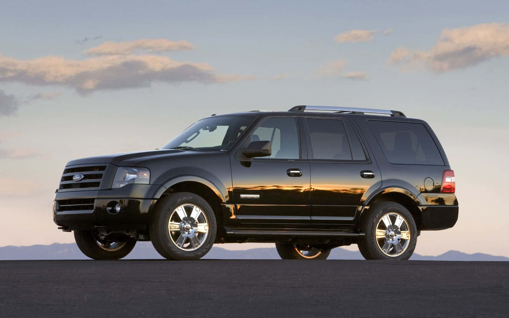 Ford Expedition 桌布 壁紙 — 一輛時尚的黑色福特 Expedition SUV 停在深色瀝青路面上，背景是柔和的黃昏天空，遠處有山脈和散落的雲朵。