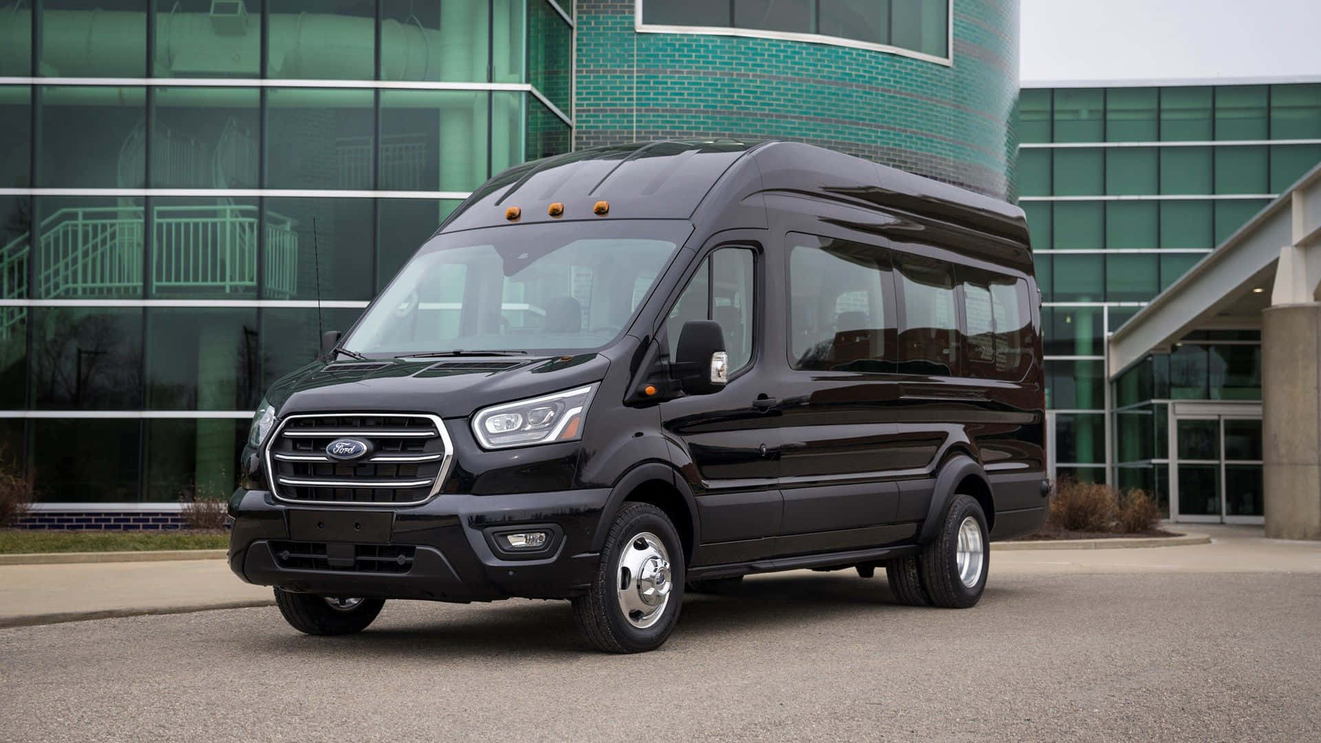 Ford Transit 桌布 壁紙 — 一輛時尚的黑色福特 Transit 停在一棟現代玻璃和磚砌建築前，天氣陰沉。