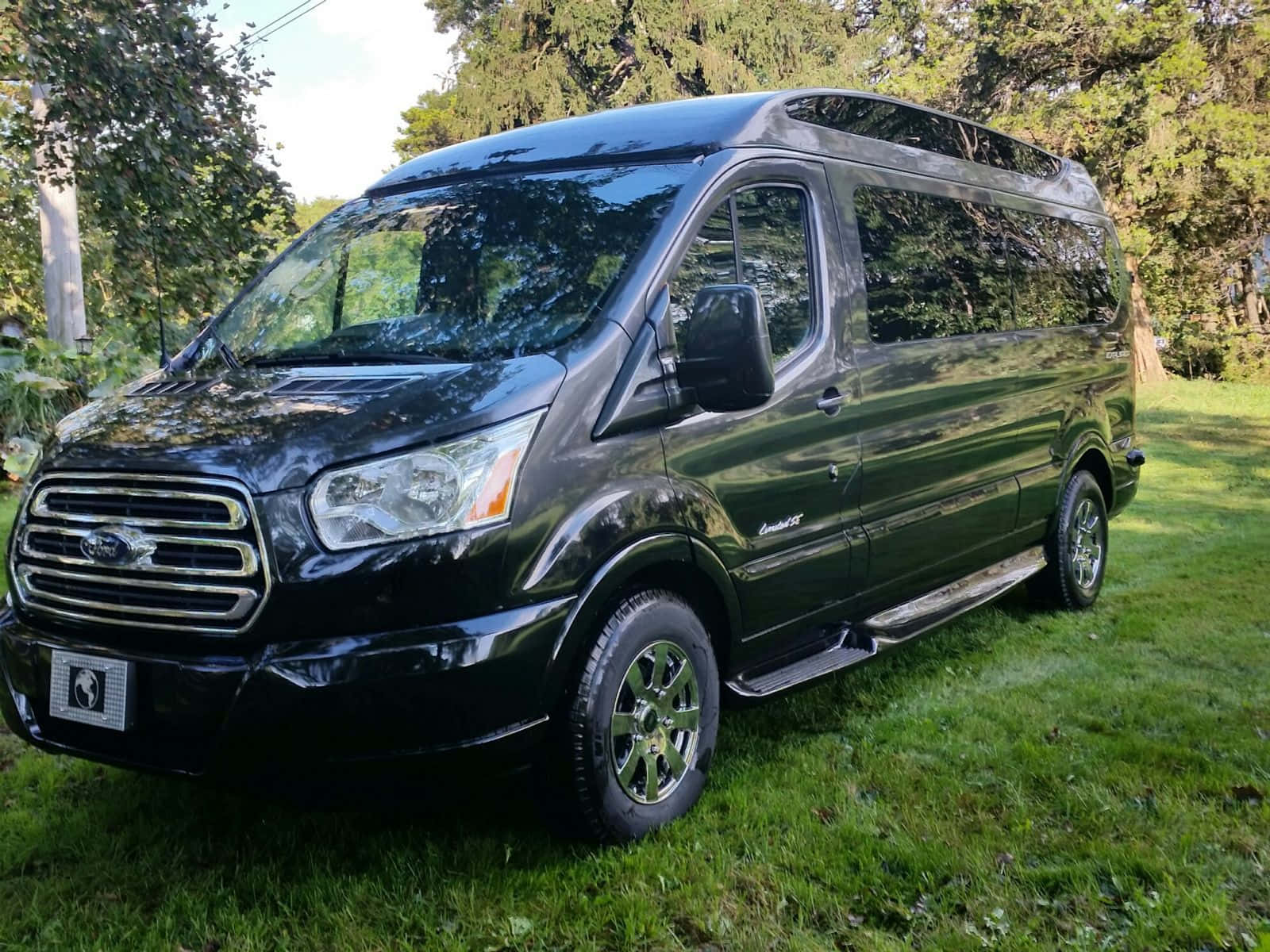 Ford Transit 桌布 壁紙 — 一輛時尚的黑色福特 Transit 客貨車，帶有鍍鉻飾邊，在晴朗的日子停泊在綠草如茵的樹蔭下。