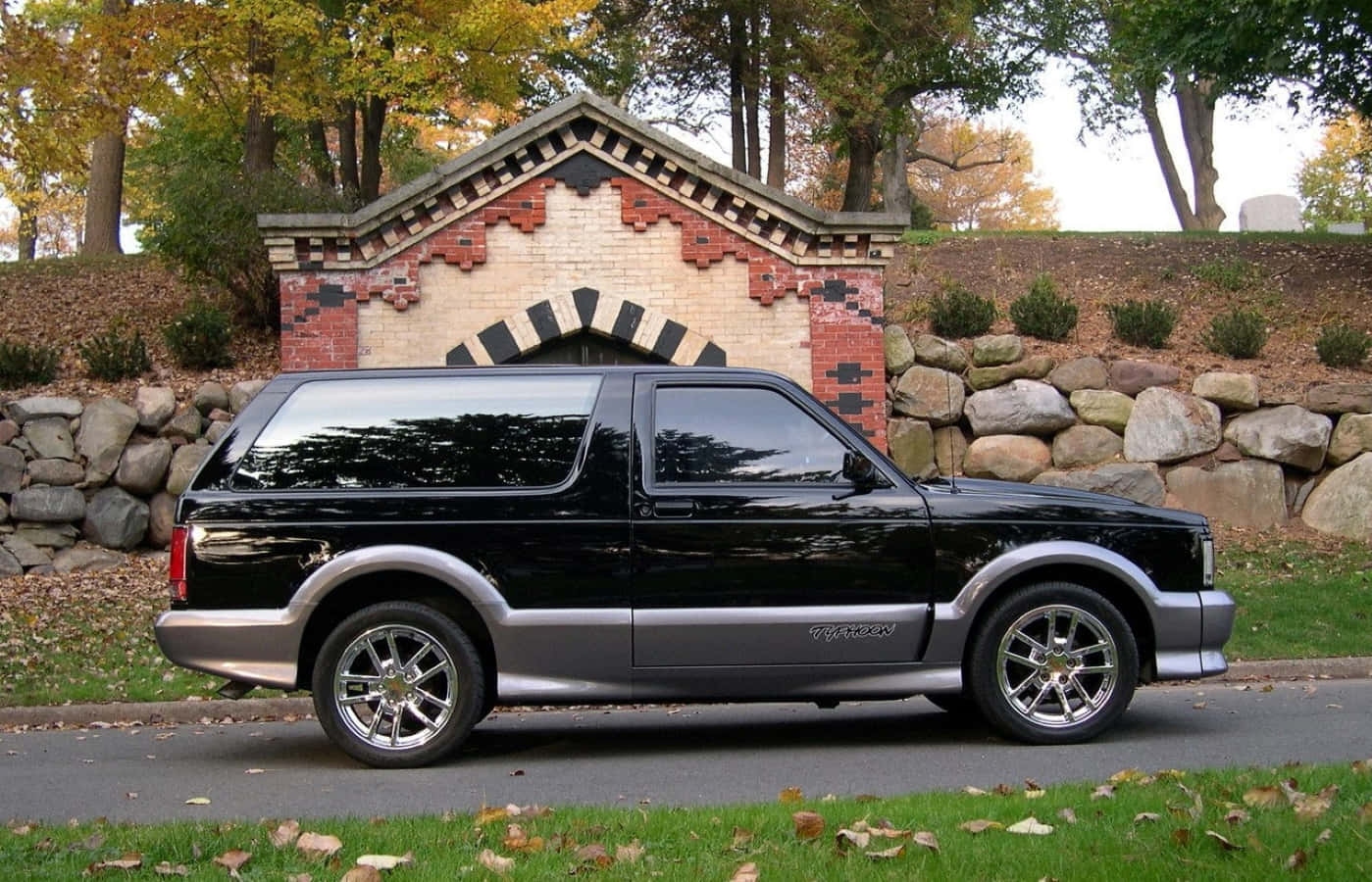 Gmc Typhoon 桌布 壁紙 — 一輛時尚的黑色福特探險者SUV，帶有鍍鉻裝飾，停在鋪砌的道路上，旁邊是草地，有落葉，背景是裝飾性磚拱門。