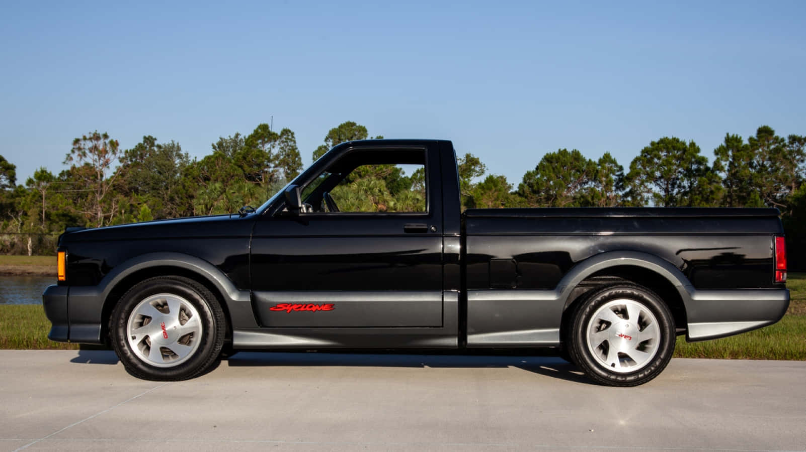 Gmc Syclone 桌布 壁紙 — 一輛時尚的黑色福特皮卡車，帶有紅色 'Syclop' 貼紙，停在柏油路上，旁邊是樹木成行的水道，上方是清澈的藍天。