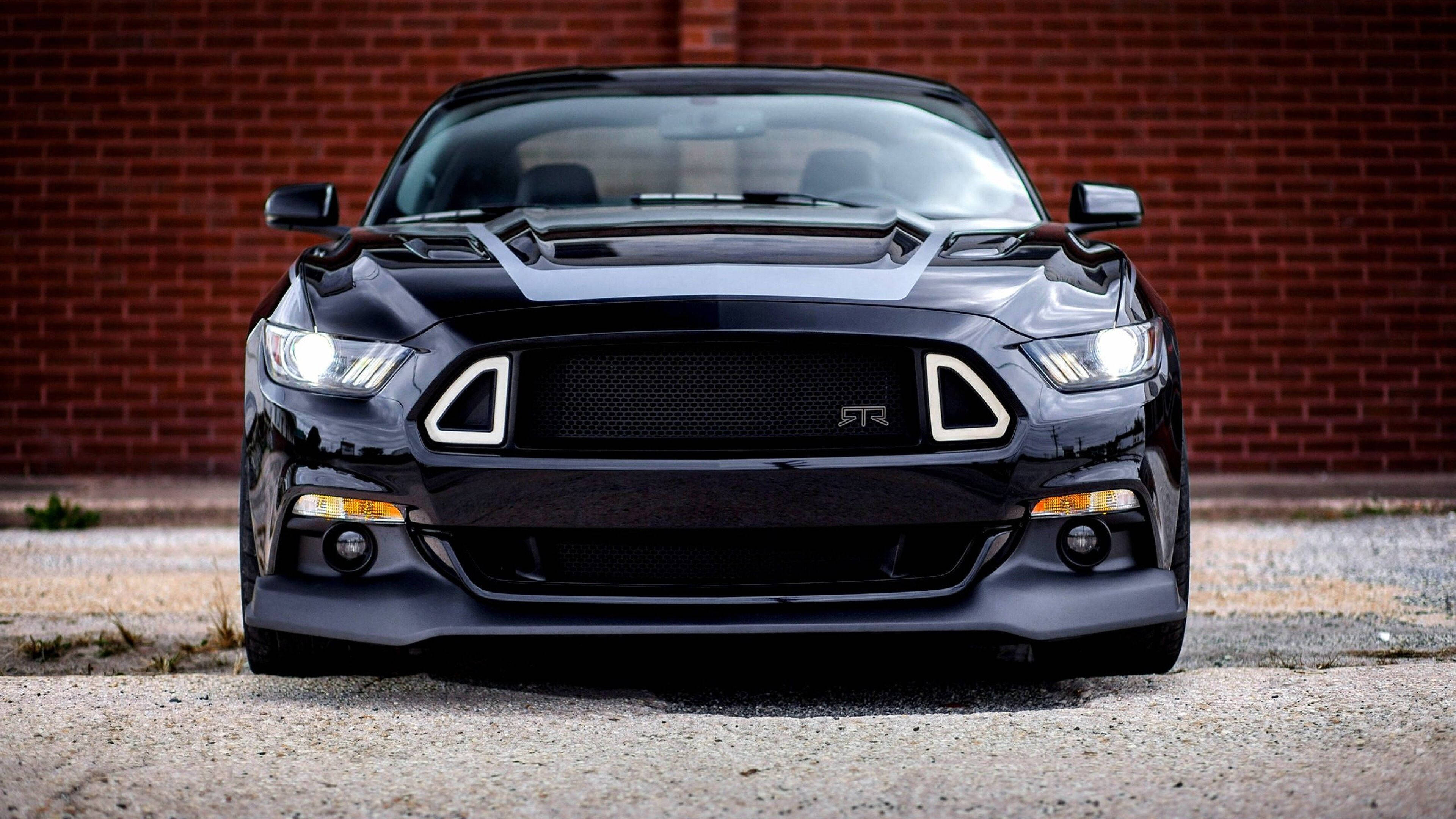 mustang HD 桌布 壁紙 — 一輛時尚的黑色福特野馬GT350R，具有侵略性造型和發光頭燈，停在風化的磚牆前。