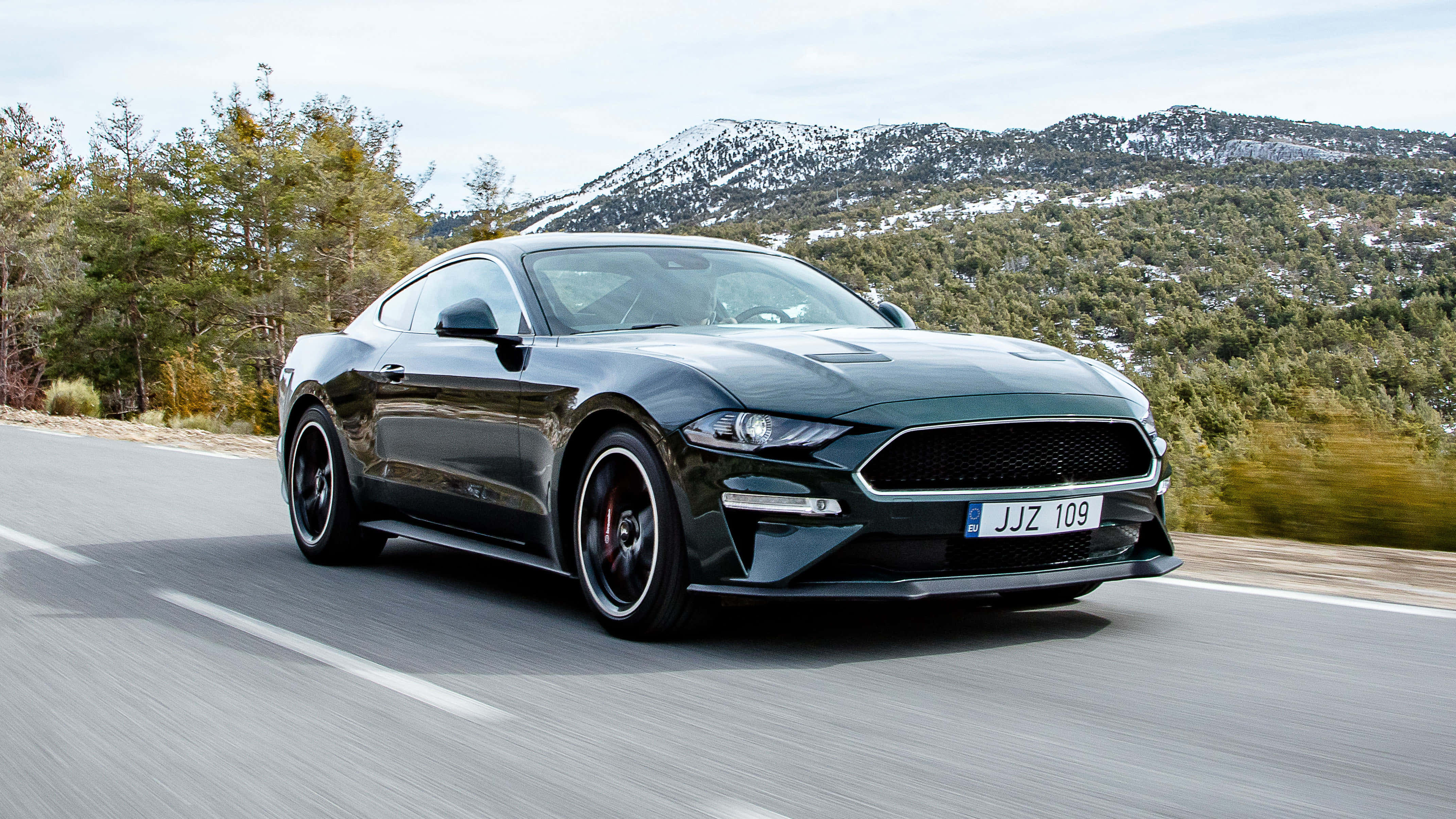 Ford Mustang Bullitt 桌布 壁紙 — 一輛時尚的黑色福特野馬在蜿蜒的山路上疾馳，背景是白雪覆蓋的山峰和松樹。