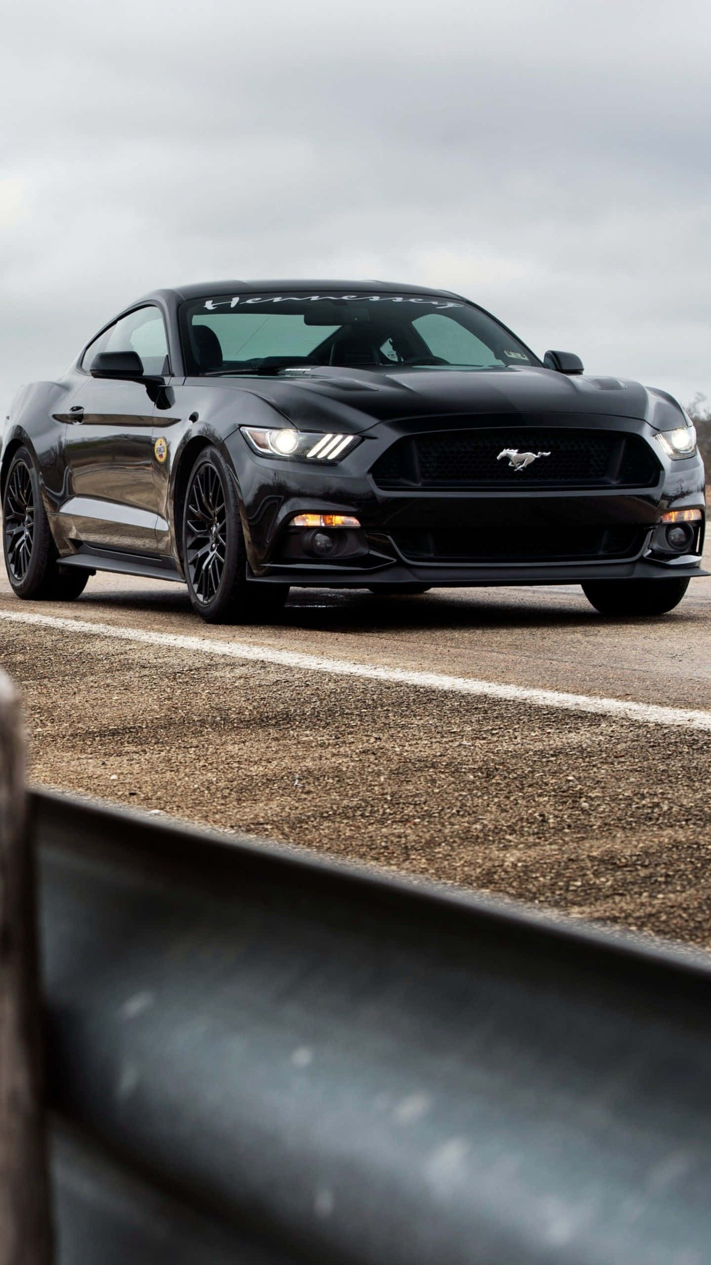 Mustang iPhone 桌布 壁紙 — 一輛時尚的黑色福特野馬跑車在陰天下行駛在碎石路上，前景有模糊的護欄。