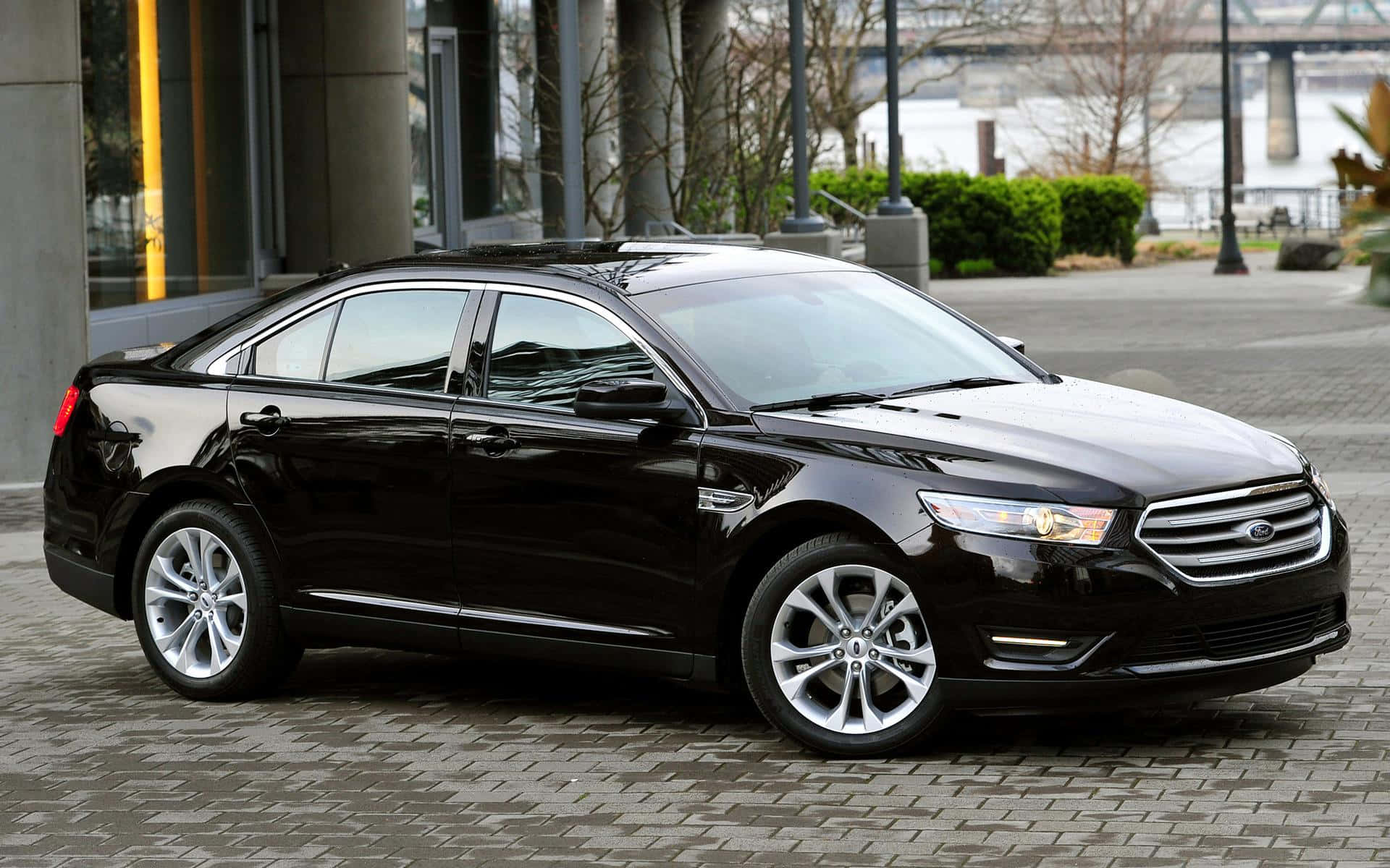 Ford Taurus 桌布 壁紙 — 一輛時尚的黑色福特金牛座轎車停放在鋪砌的城市廣場上，背景是現代建築和光禿禿的樹木。