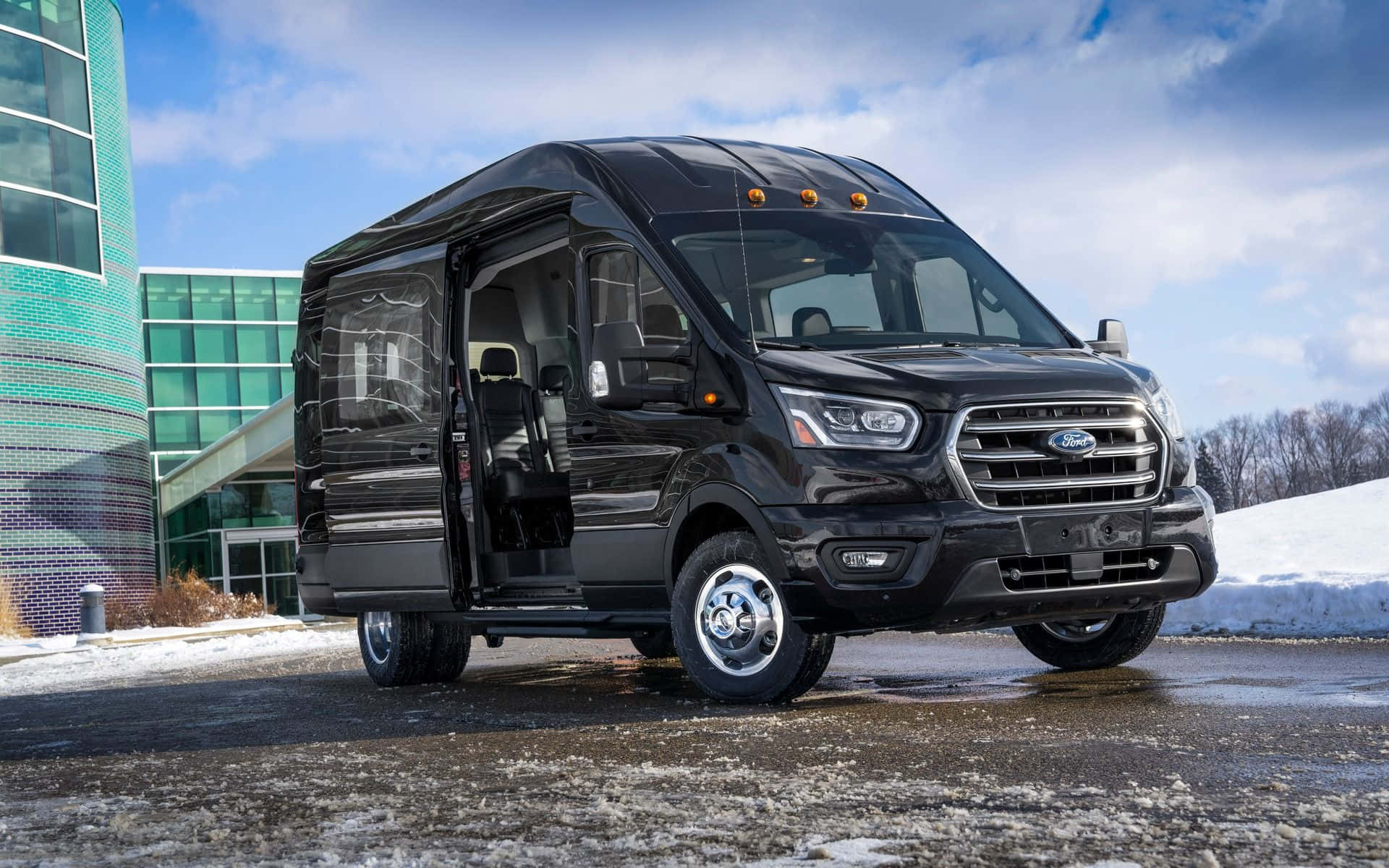Ford Transit 桌布 壁紙 — 一輛時尚的黑色福特露營車停放在雪地上，旁邊是一座現代玻璃建築，上方是明亮的藍天。