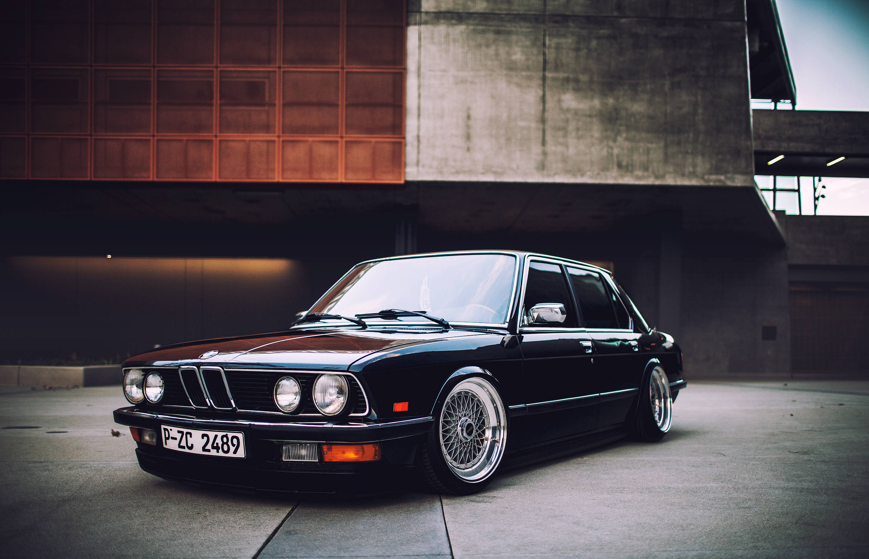 classic bmw 桌布 壁紙 — 一輛時尚的黑色經典BMW轎車配備定制輪圈，停放在現代混凝土和橙色面板建築物下。