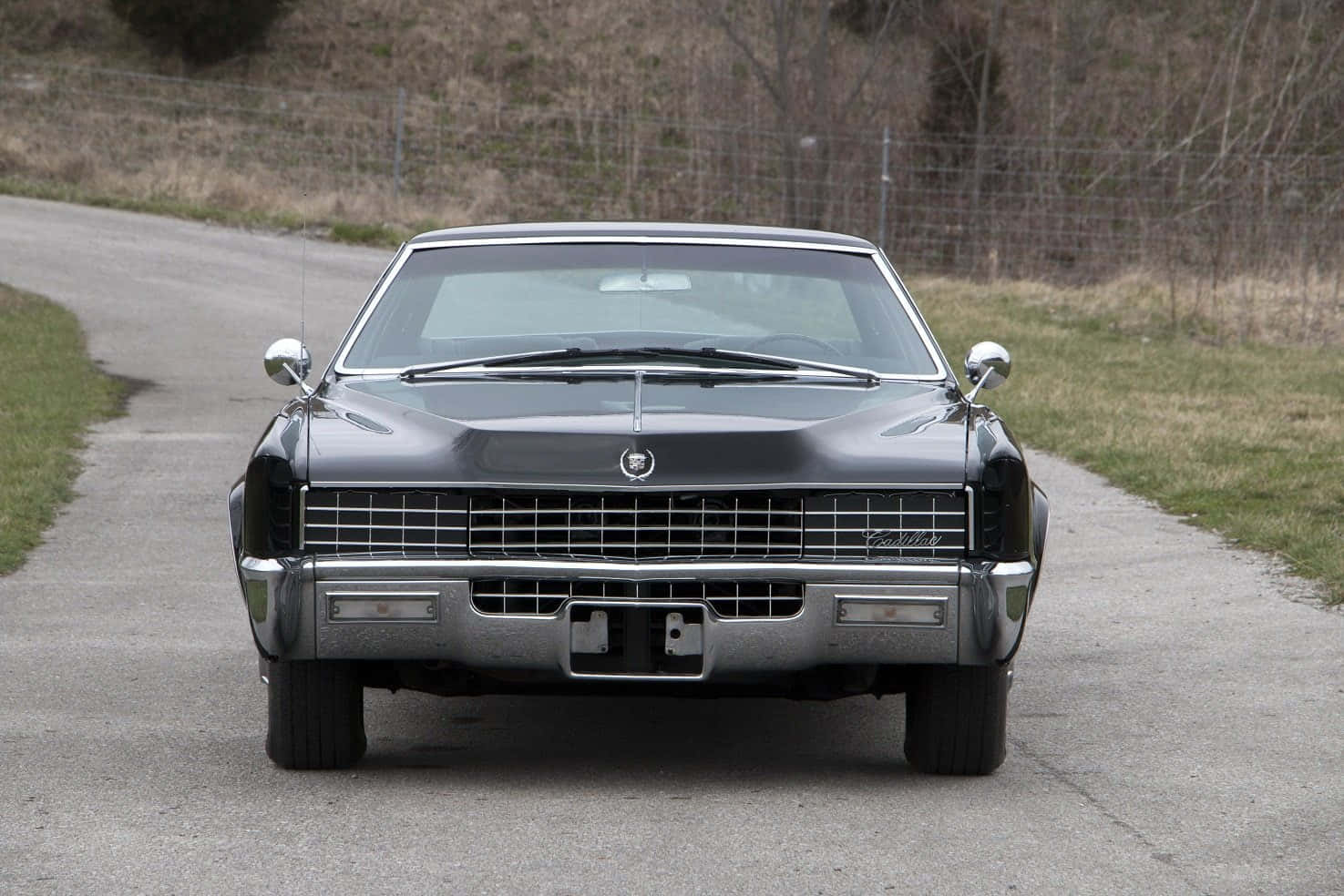 Cadillac Fleetwood 桌布 壁紙 — 一輛時尚的黑色經典凱迪拉克轎車在寧靜的鄉村道路上向前行駛，兩旁是草地和光禿禿的樹木。