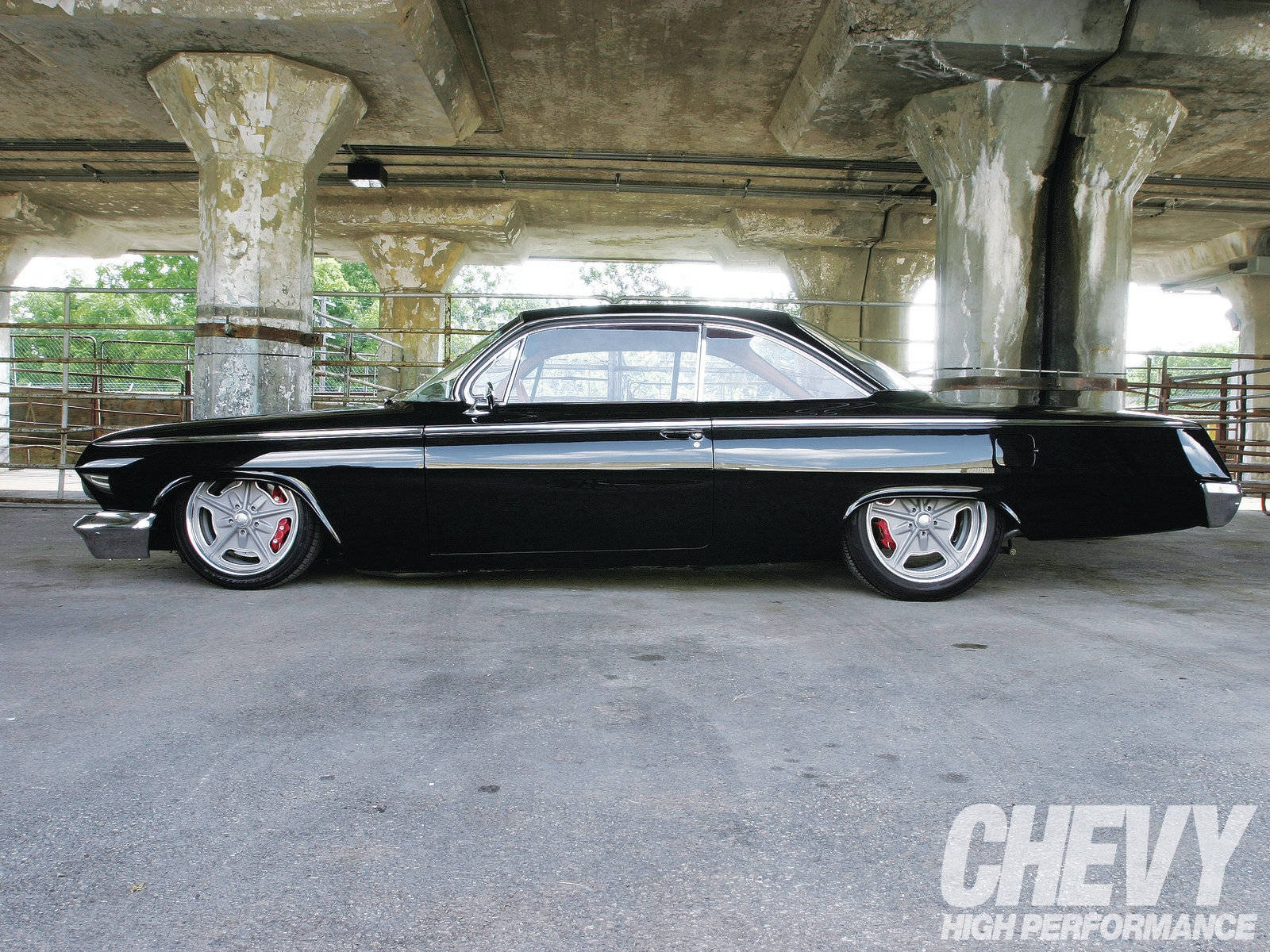 Lowrider 桌布 壁紙 — 一輛時尚的黑色經典肌肉車，帶有鍍鉻裝飾和紅色制動卡鉗，停在風化的混凝土天橋下。