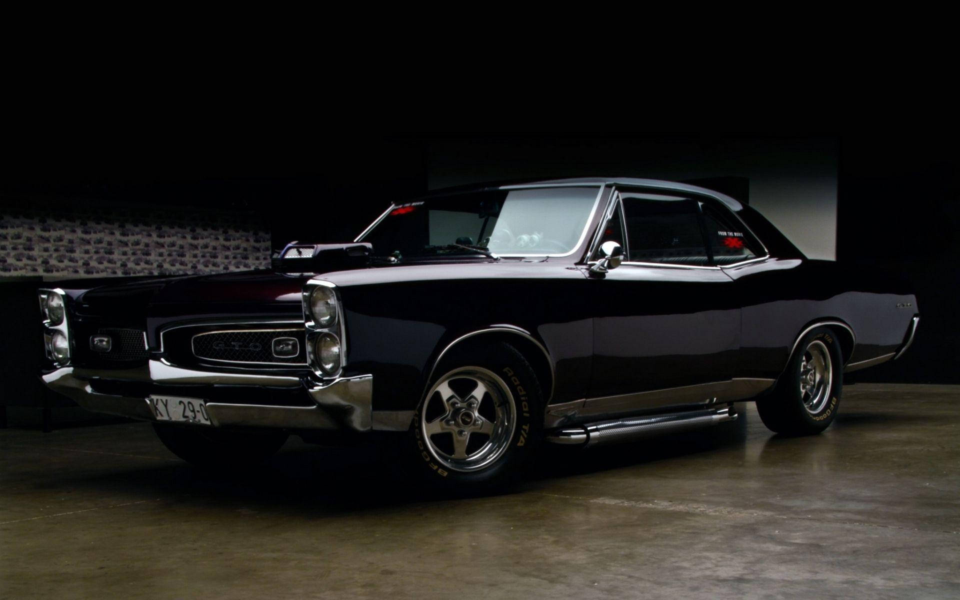 Pontiac 桌布 壁紙 — 一輛時尚的黑色經典肌肉車，帶有鍍鉻裝飾和定制輪圈，在黑暗的車庫中被戲劇性地照亮。