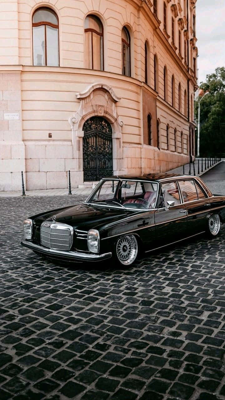 Mercedes 桌布 壁紙 — 一輛時尚的黑色經典賓士轎車停泊在鵝卵石街道上，旁邊是一座巨大的桃色歐洲建築。