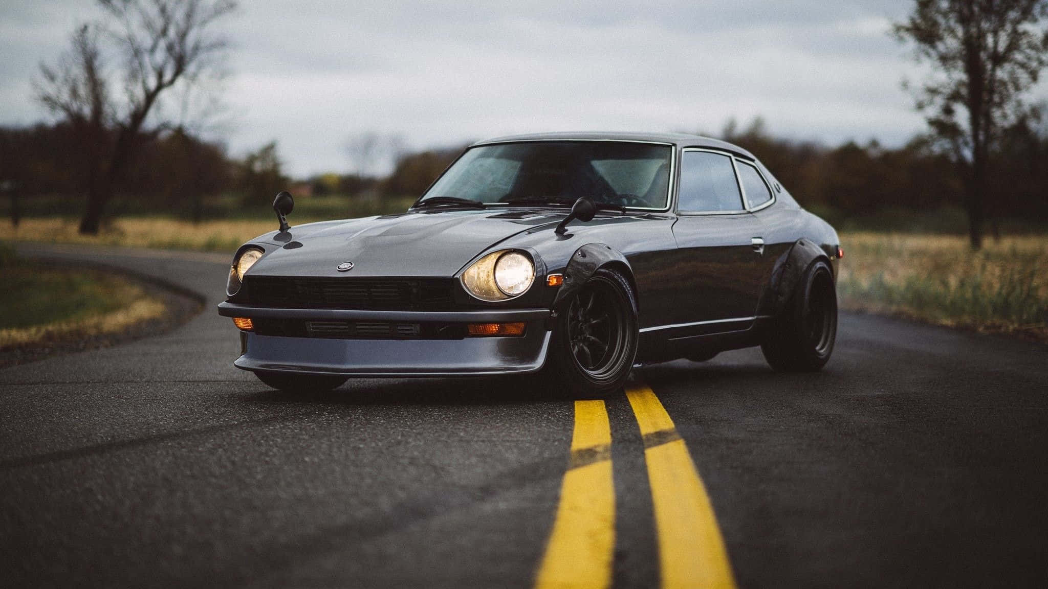 Datsun 桌布 壁紙 — 一輛時尚的黑色經典跑車，車頭燈閃爍，在陰天下行駛在一條兩旁光禿樹木的潮濕鄉村道路上。