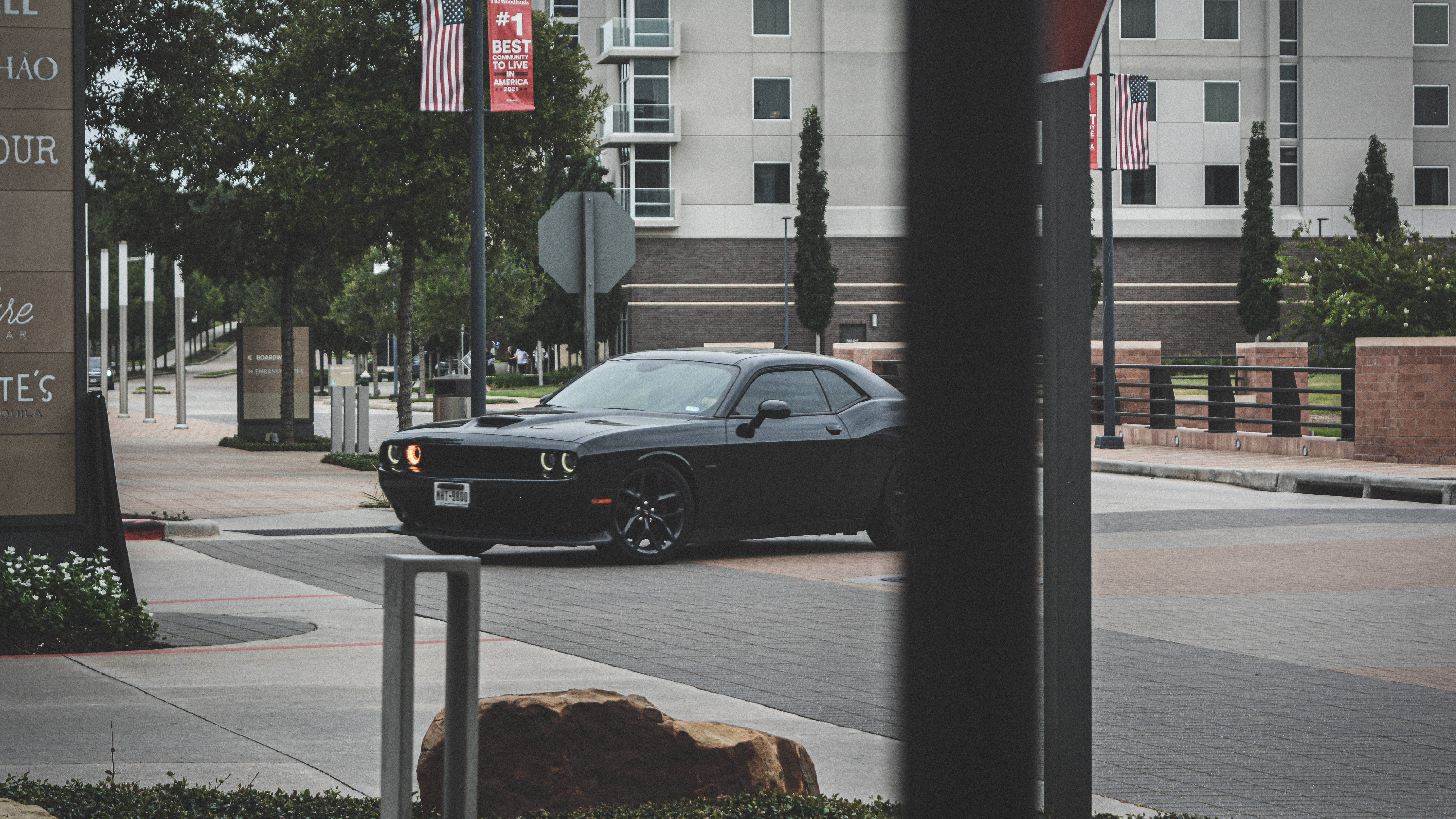 Challenger 桌布 壁紙 — 一輛時尚的黑色肌肉車行駛在現代城市街道上，前景有柱子和磚砌景觀。
