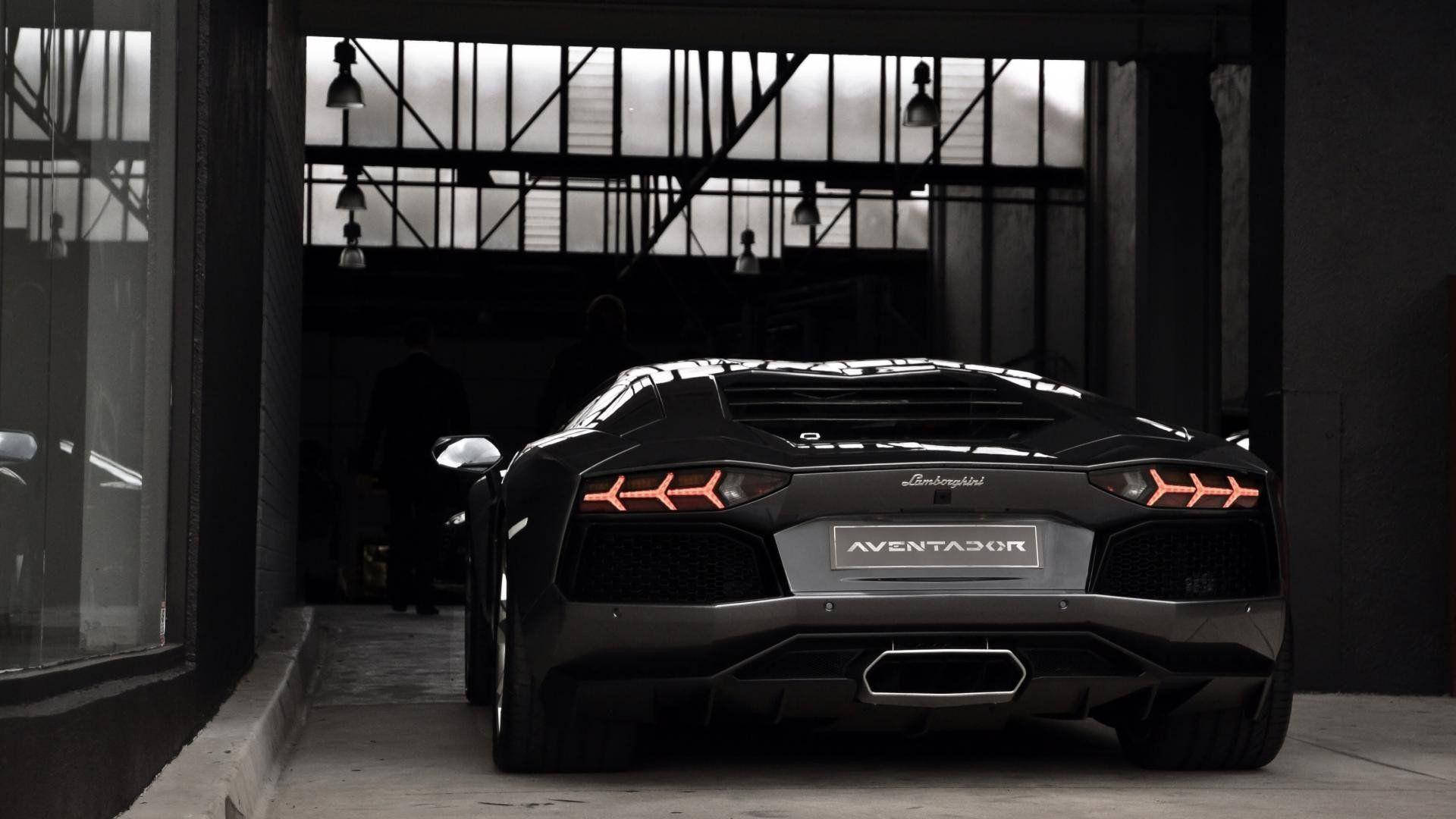 Lamborghini 4K 桌布 壁紙 — 一輛時尚的黑色藍寶堅尼Aventador，帶有發光的紅色尾燈，停放在昏暗的工業車庫內。