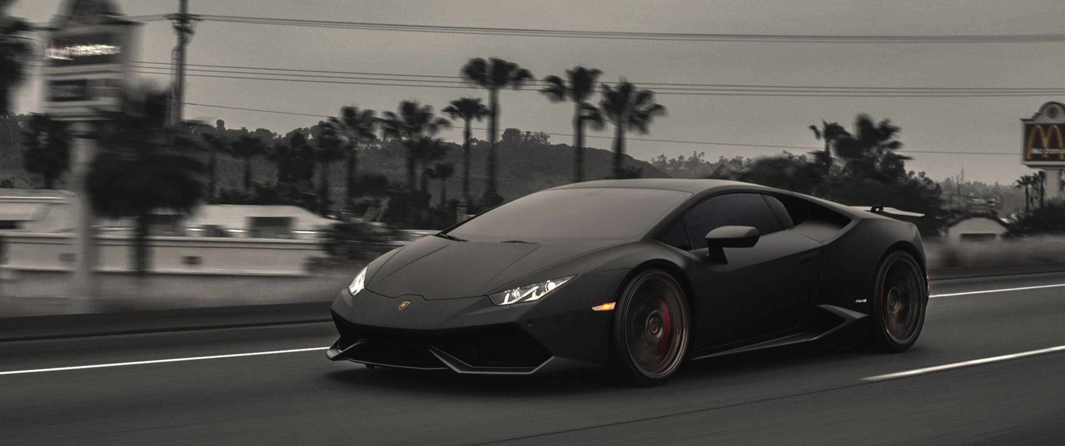 3440x1440p Lamborghini 桌布 壁紙 — 一輛時尚的黑色藍寶堅尼Huracán在棕櫚樹成蔭的街道上奔馳，帶有動態模糊和柔和的灰色天空。