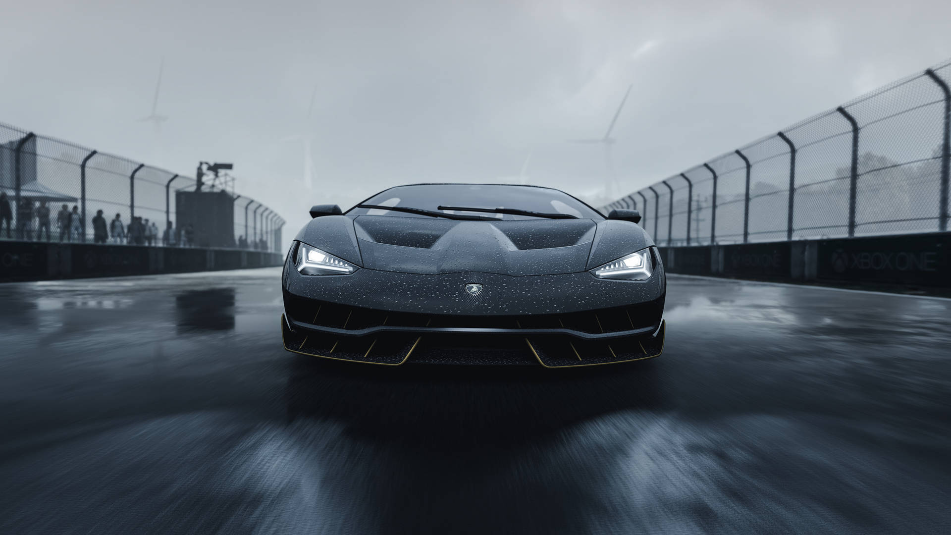 Motorsport 桌布 壁紙 — 一輛時尚的黑色藍寶堅尼在潮濕的賽道上，在陰天和遠處的風力發電機背景下，向觀眾疾馳而來。