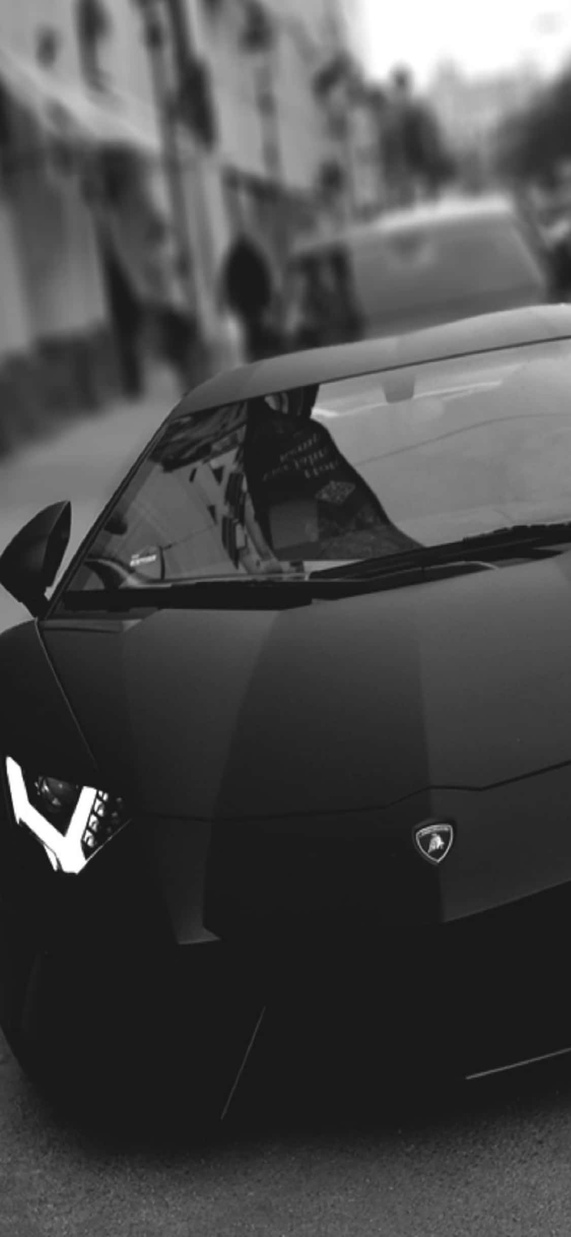iPhone X Lamborghini 桌布 壁紙 — 一輛時尚的黑色藍寶堅尼跑車，帶有發光的車頭燈，停放在單色模糊的城市街道上。