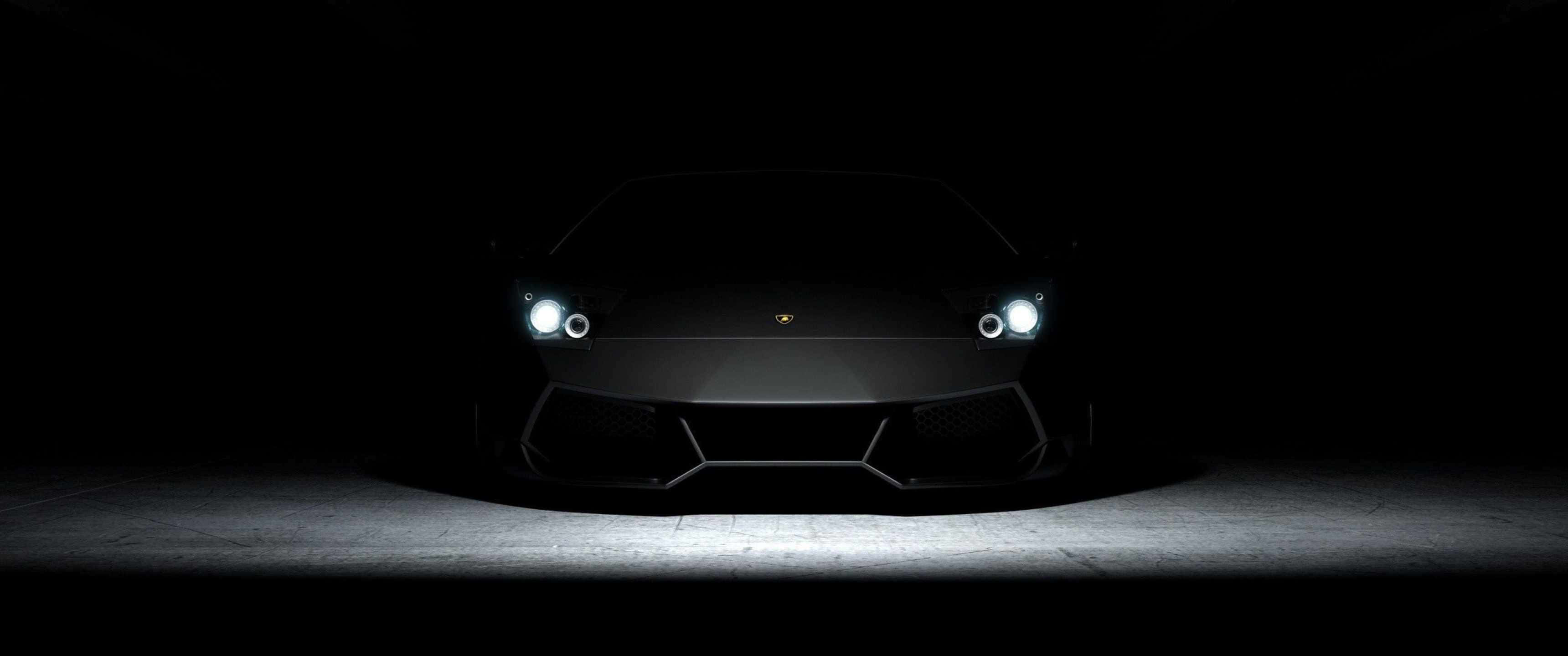 3440x1440p Lamborghini 桌布 壁紙 — 一輛時尚的黑色藍寶堅尼跑車在漆黑一片的虛空中閃爍著刺眼的車頭燈，聚光燈照射在混凝土車道上。