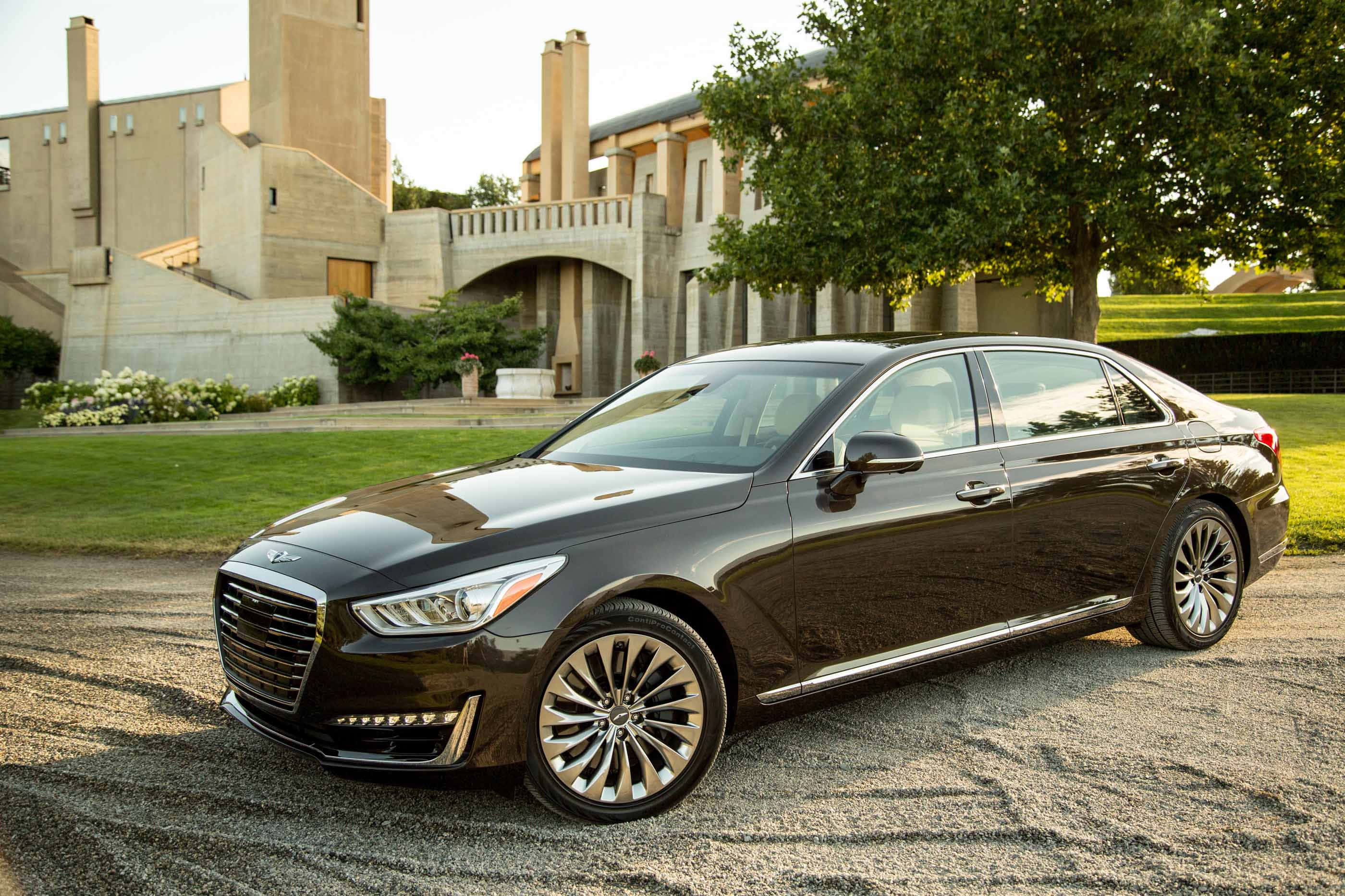 Genesis G90 桌布 壁紙 — 一輛時尚的黑色豪華轎車停在碎石路上，前方是一座現代石建築，周圍有茂密的綠樹和修剪整齊的草坪。