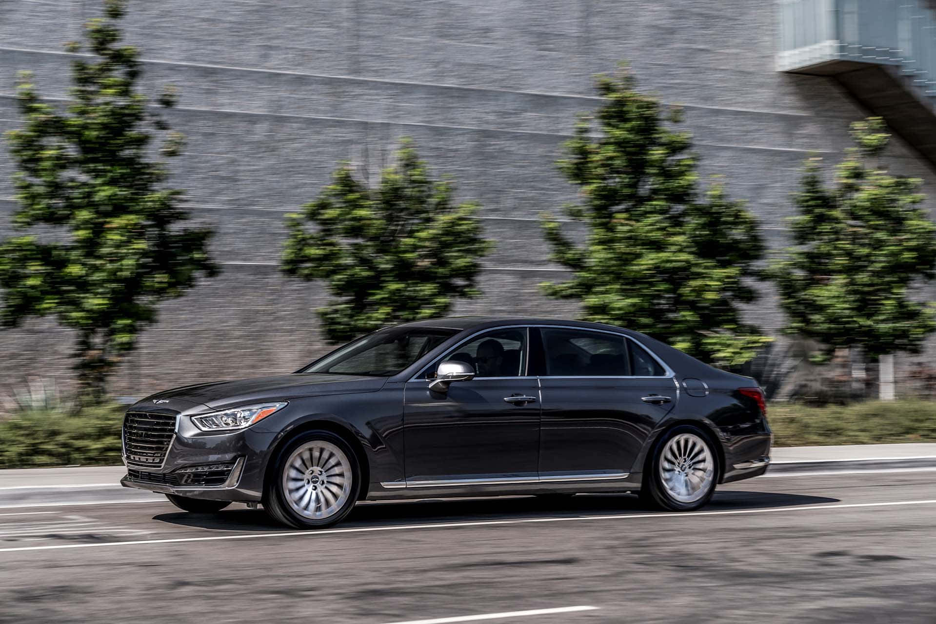 Genesis G90 桌布 壁紙 — 一輛時尚的黑色豪華轎車在城市的街道上飛馳，背景是模糊的綠色樹木和灰色混凝土牆。