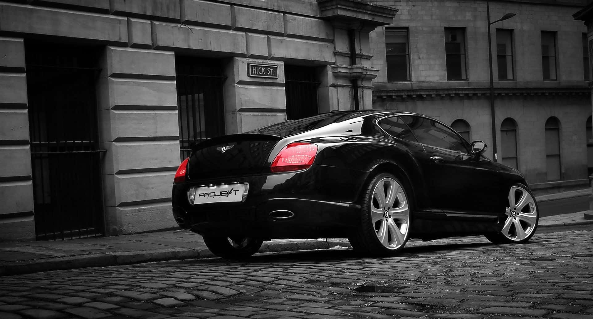 Best Bentley 桌布 壁紙 — 一輛時尚的黑色賓利 Continental GT 停在鵝卵石街道上，在單色城市背景下，紅色尾燈顏色選擇性彈出。